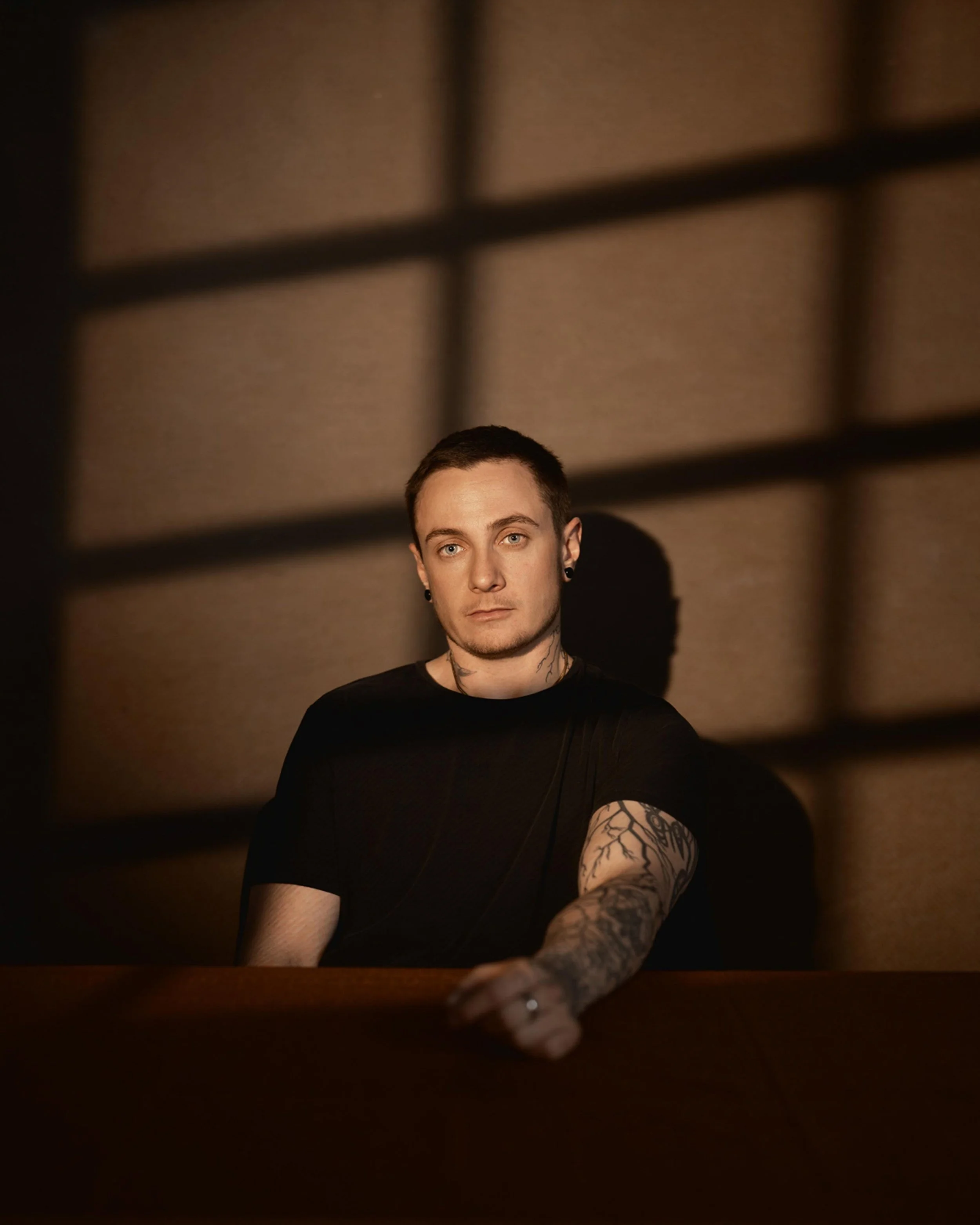 A young man with short dark hair and tattoos on his arms and neck, wearing a black shirt, sitting at a dark wooden table with a serious expression, in a dimly lit room with overhead grid-patterned lighting.
