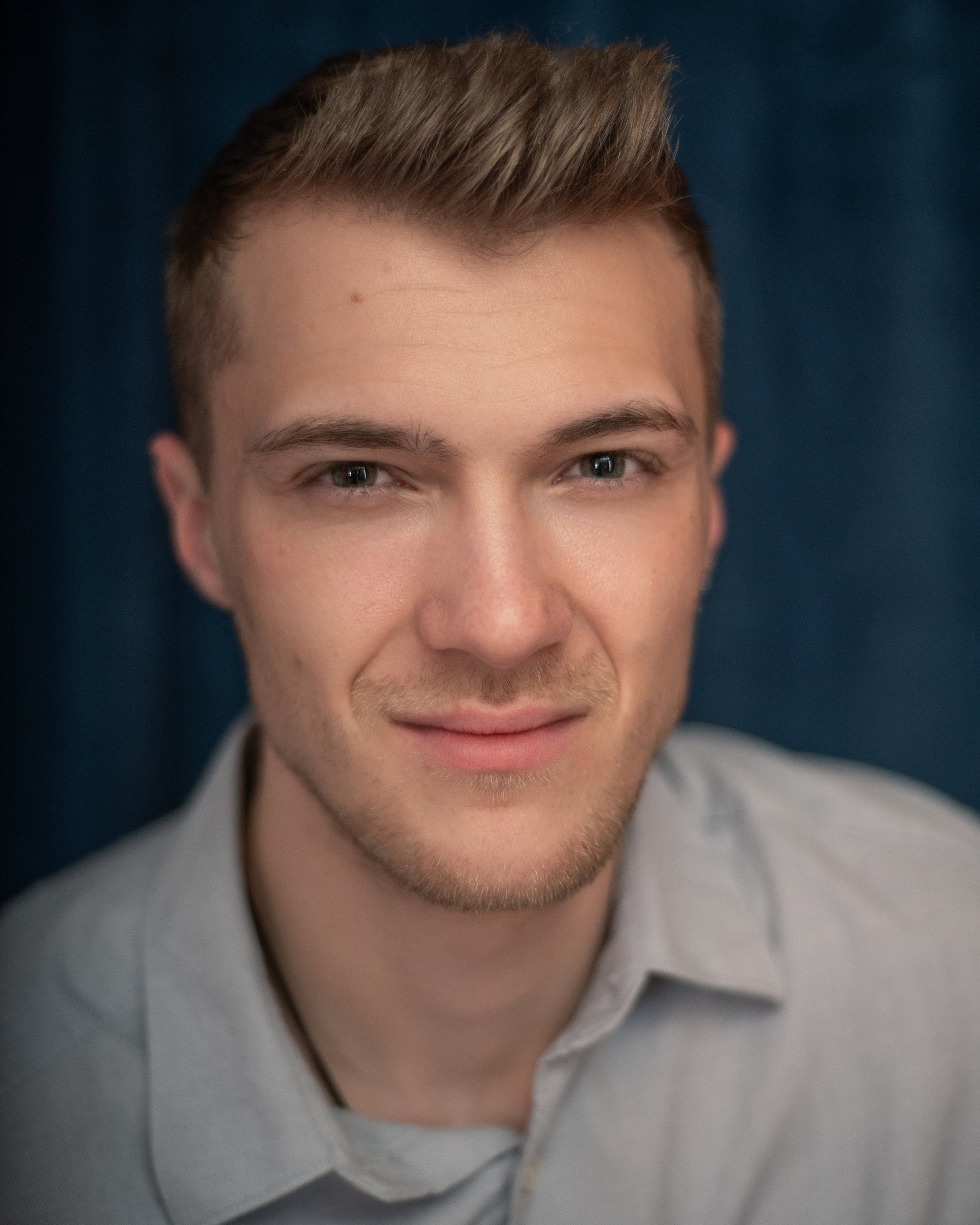 Headshot of a man with blond hair and a light grey shirt in front of a blue backdrop
