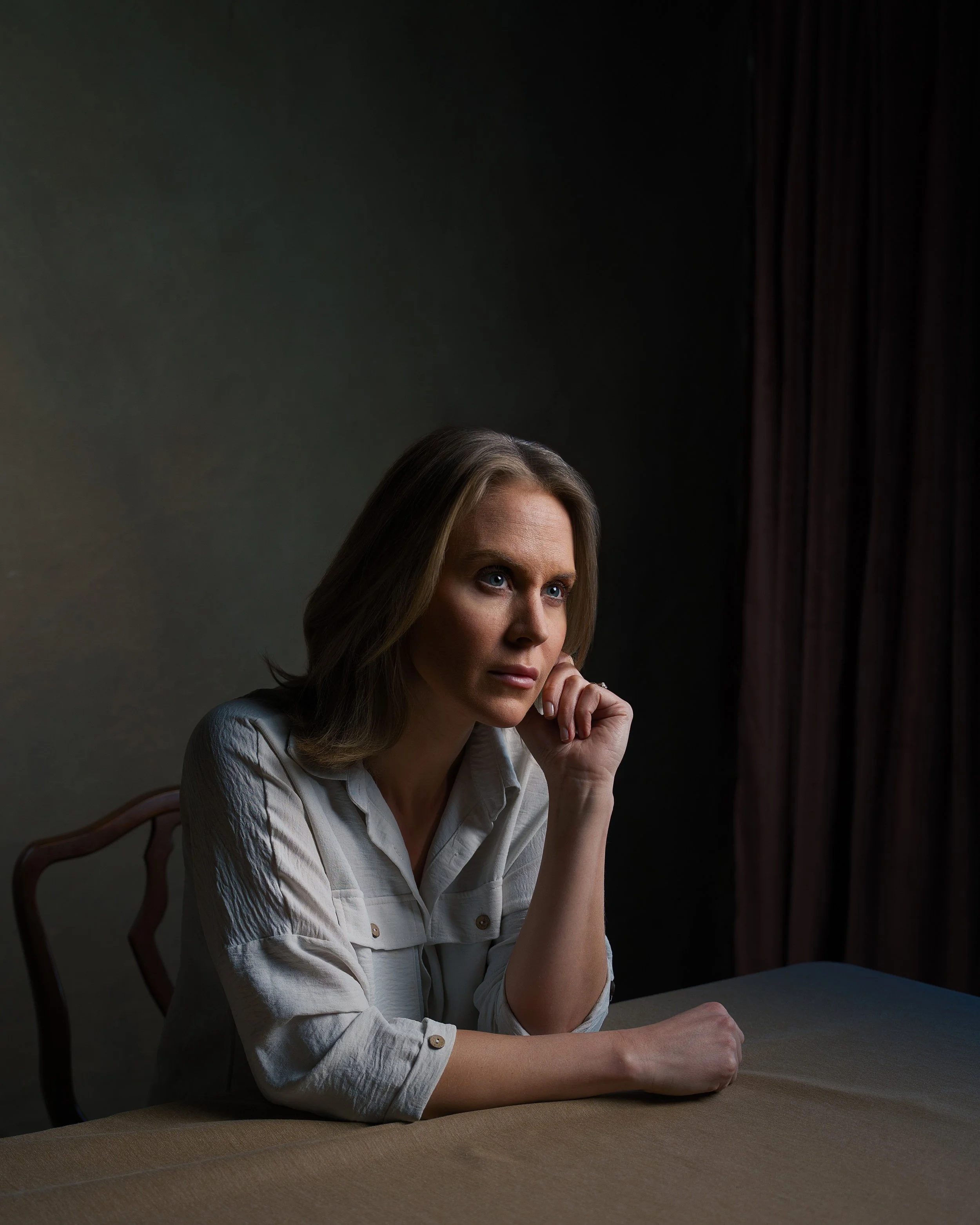 A woman with brown hair and blue eyes sitting at a wooden table, resting her chin on her hand, with a dark background and red curtain on the side.