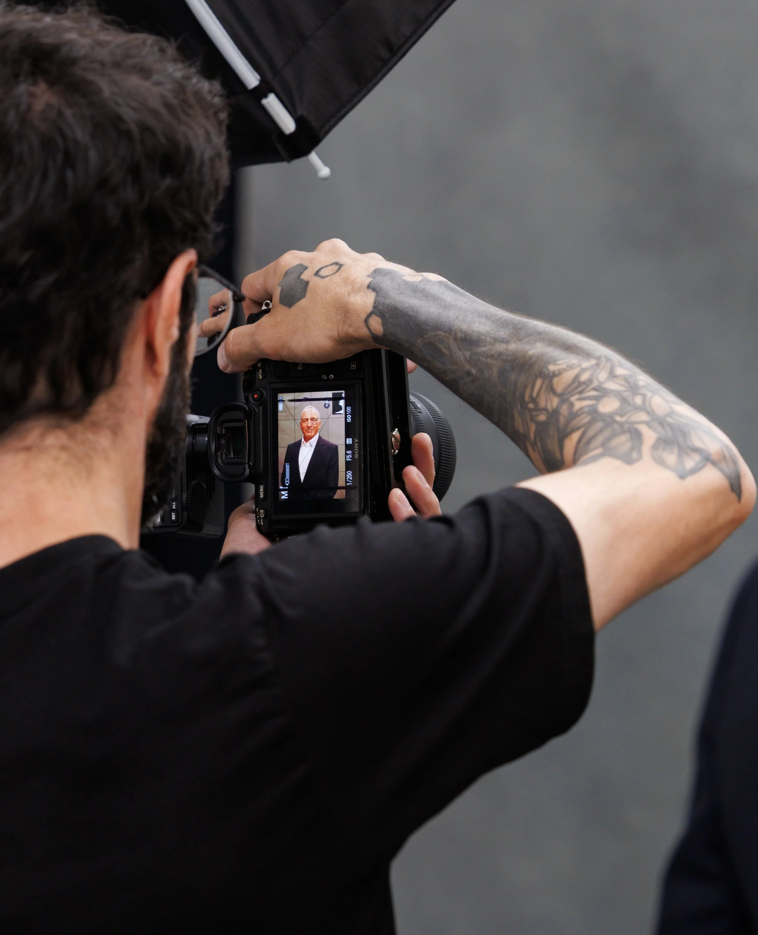A person with tattoos on their arm is adjusting settings on a camera, with the camera screen displaying a portrait of an older man in a suit and tie.
