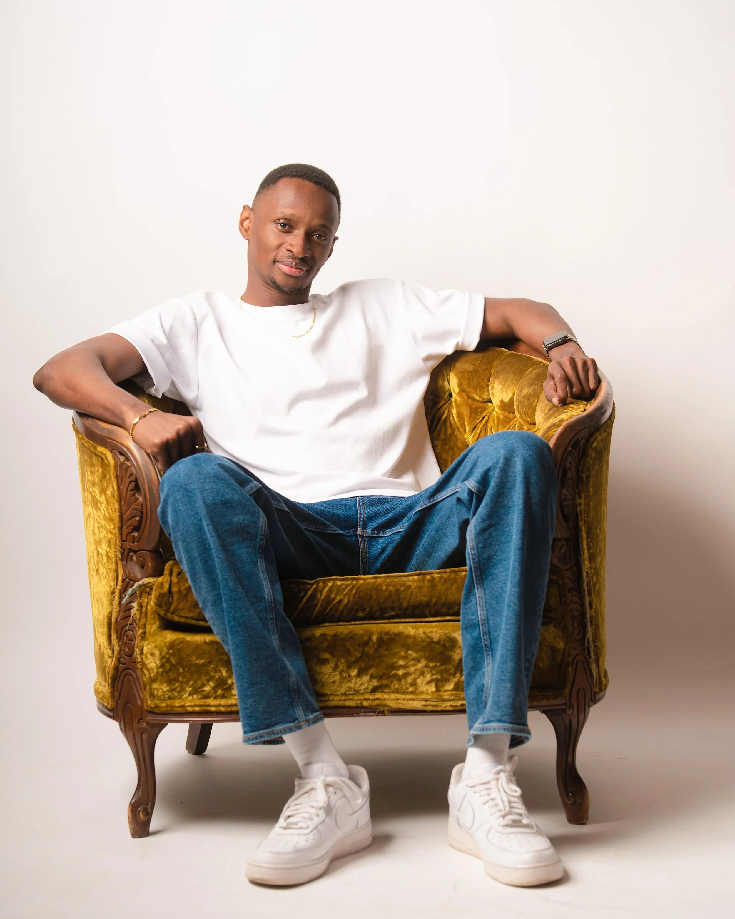 A man with short hair wearing a white T-shirt, blue jeans, and white sneakers, sitting in a mustard velvet vintage armchair with wooden carved legs, against a plain white wall.
