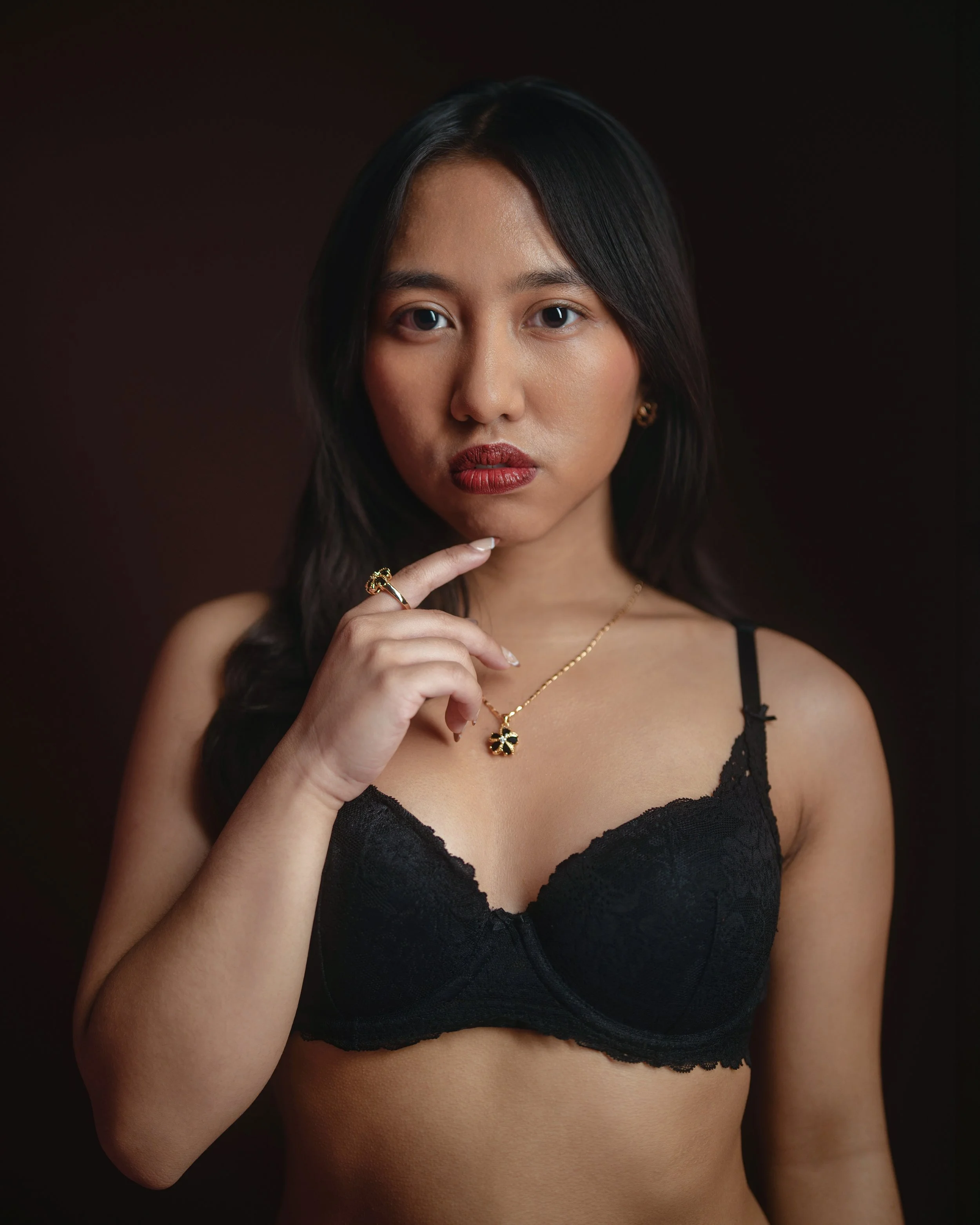 A young woman with black hair and medium skin tone wearing a black lace bralette and gold jewelry, including a necklace, earrings, and rings, against a dark background.