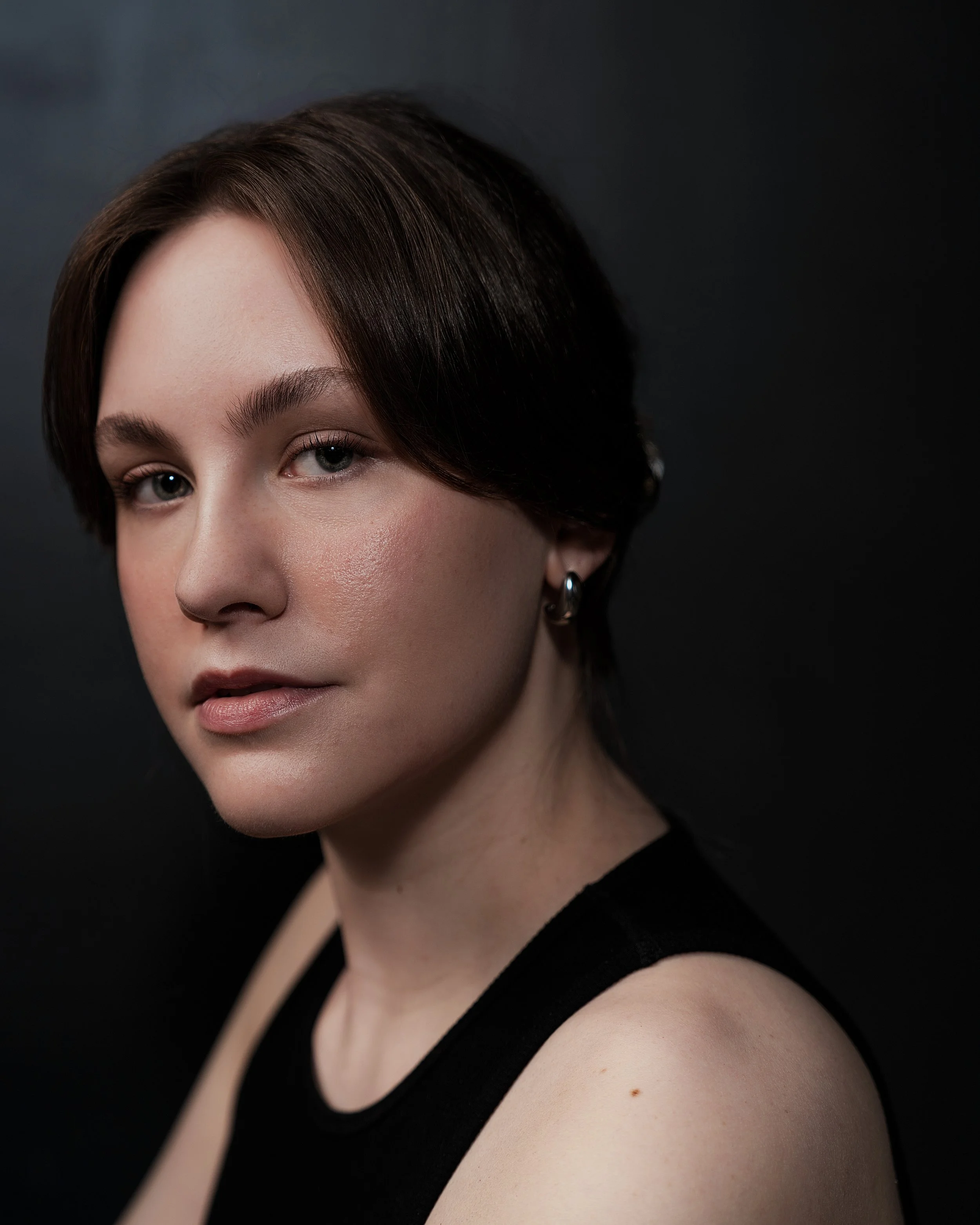 Close-up portrait of a woman with fair skin, short dark brown hair, wearing a black sleeveless top, and silver earrings, against a dark background.