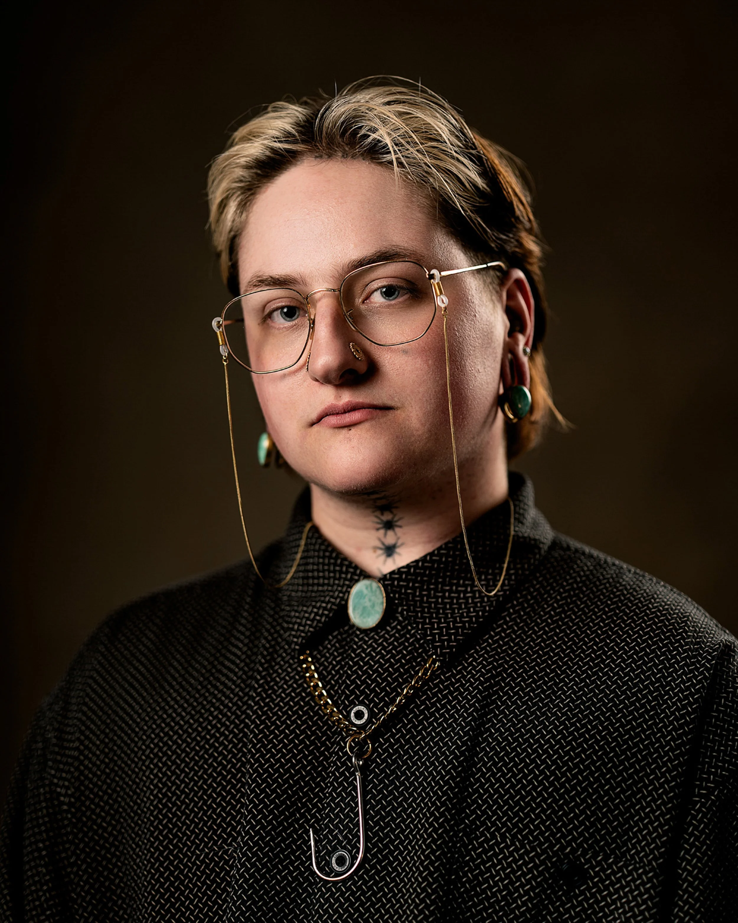 A person with short hair wearing wire glasses, earrings, and layered necklaces, dressed in a dark textured top, against a dark background.