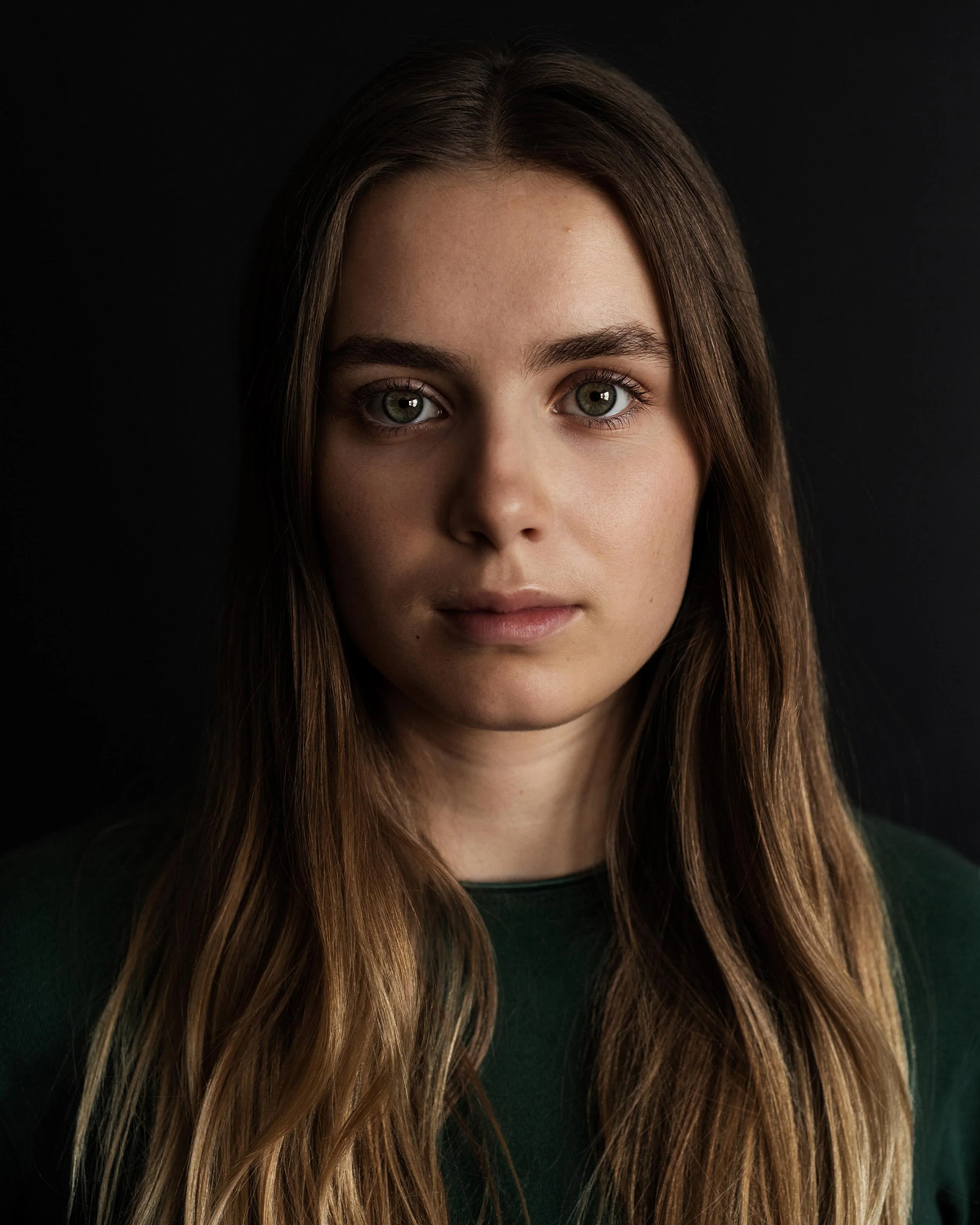 Close-up of a young woman with long brown hair and light skin, looking directly at the camera against a black background.