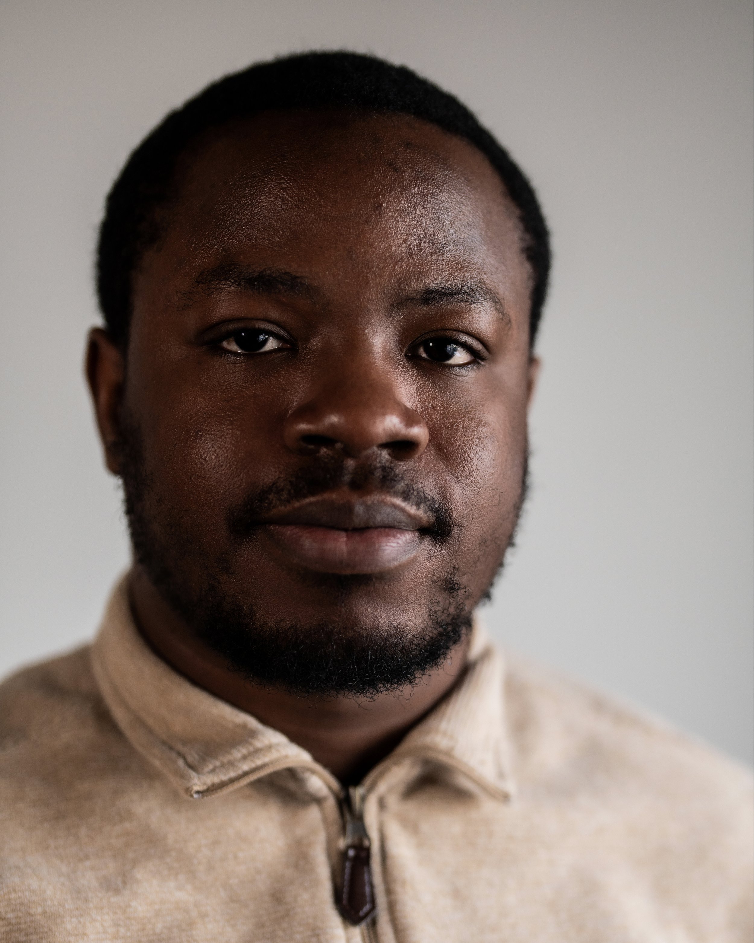 Headshot of a black man wearing a beige shirt in front of a white backdrop