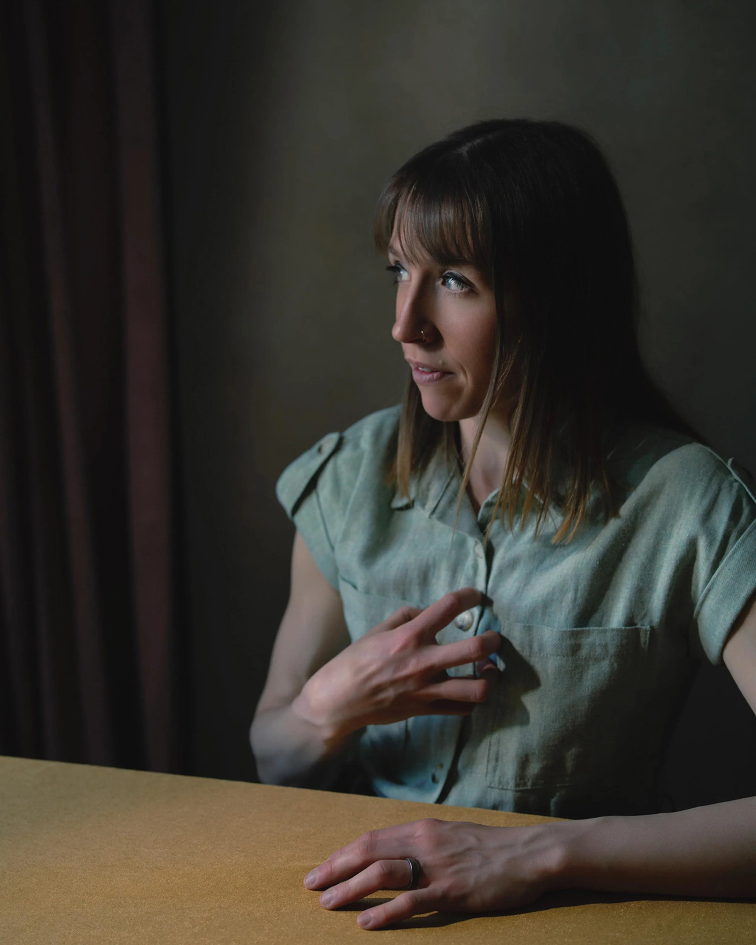 Woman with dark hair and a nose ring touches her chest, sitting at a table with a yellow surface, with her hand resting on the table in front of her.