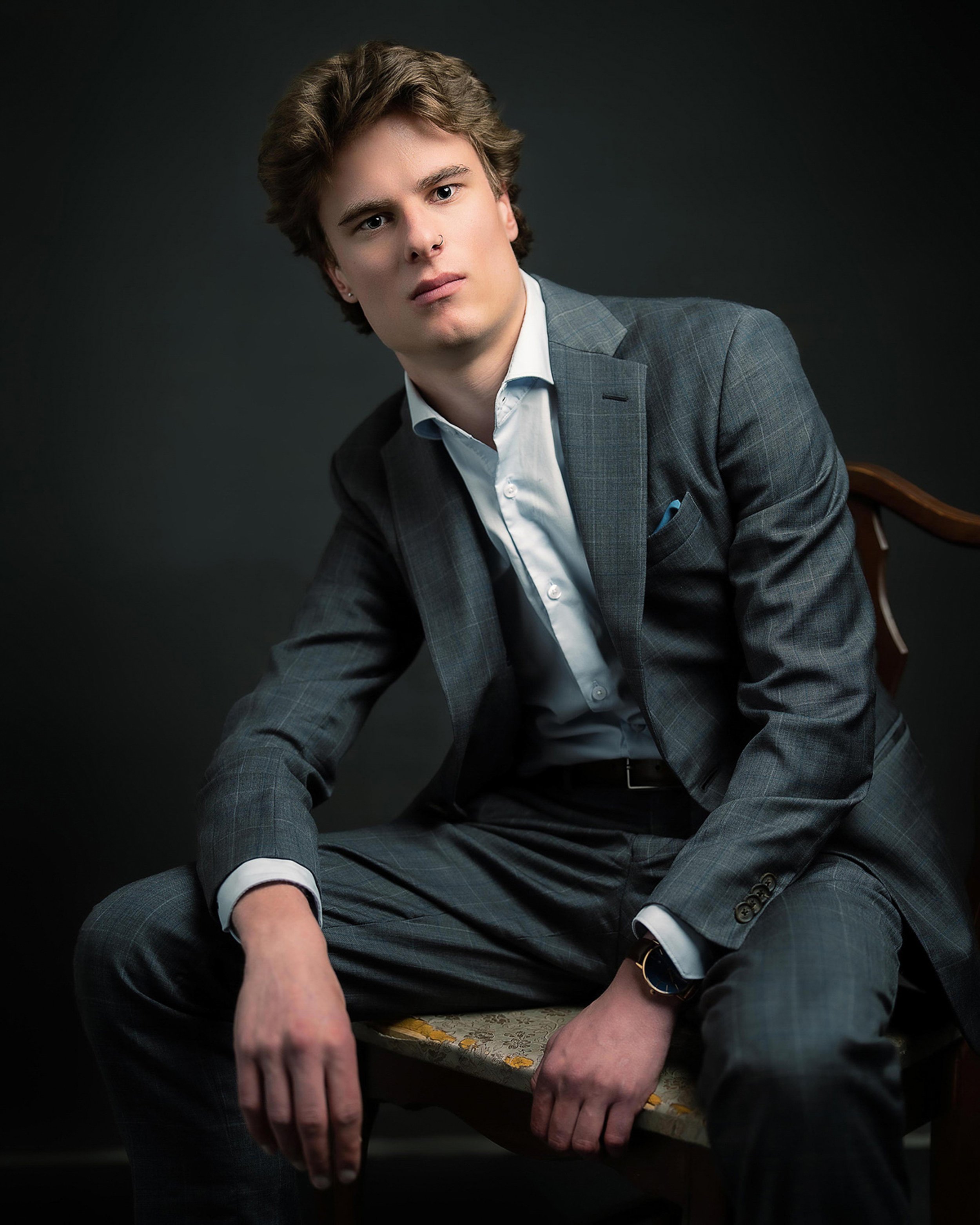 A young man with curly brown hair, wearing a grey plaid suit, white shirt, and black watch, sitting at a table with a patterned tablecloth against a dark background.
