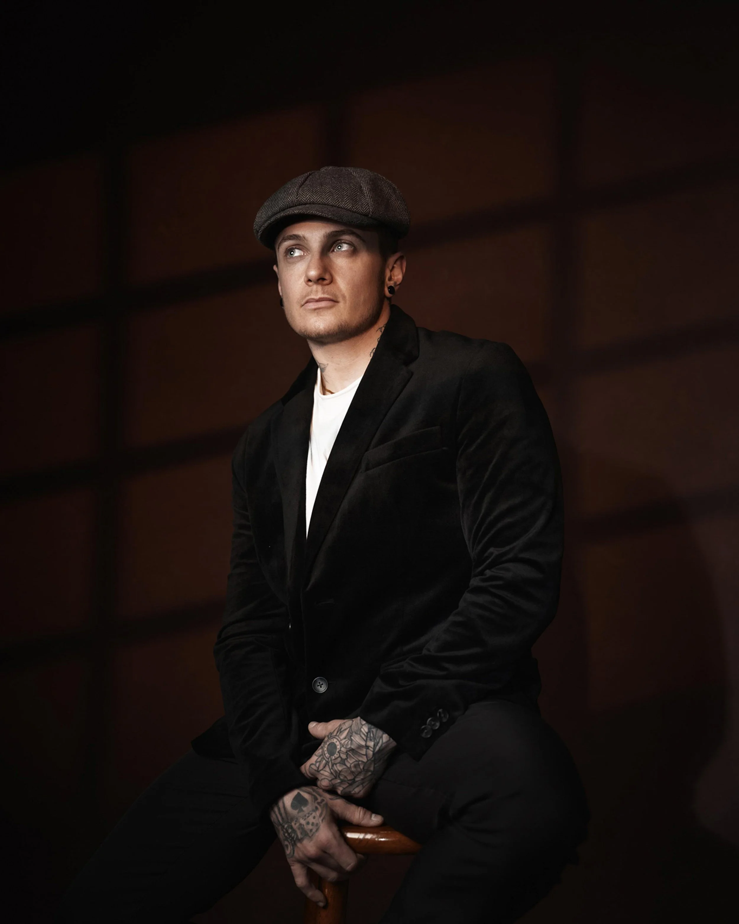 A young man with tattoos on his hands, wearing a gray flat cap, black blazer, and white shirt, sitting on a wooden stool against a dark background and looking upward to the left.