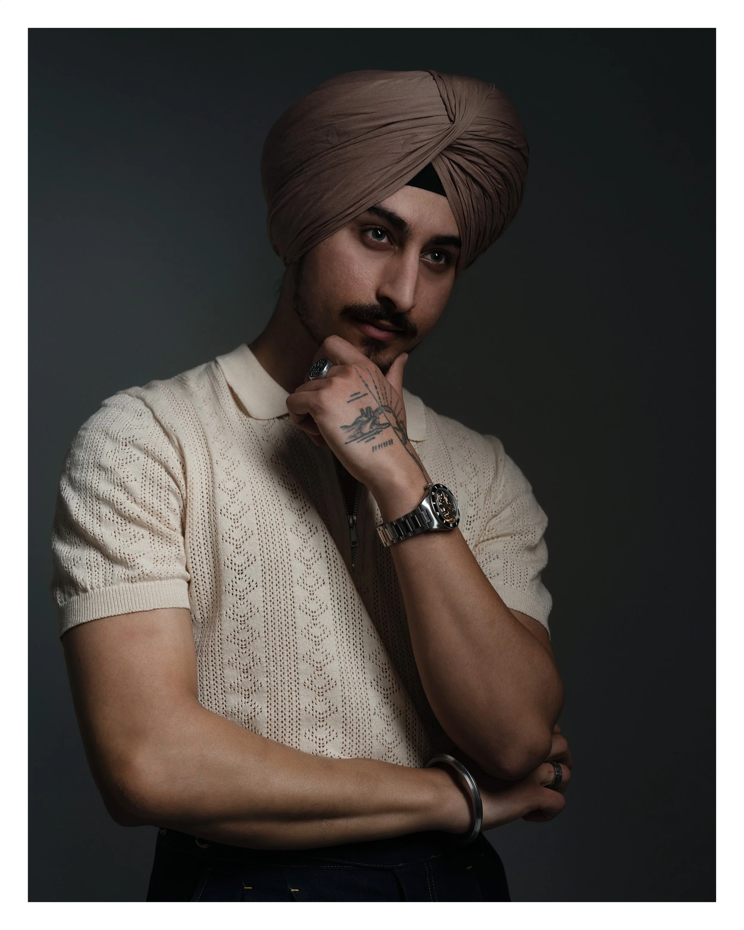 A young man with a mustache and beard wears a brown turban, a cream-colored knit sweater, and jewelry including a watch and rings. He has tattoos on his left hand and is posing with his hand under his chin against a dark background.