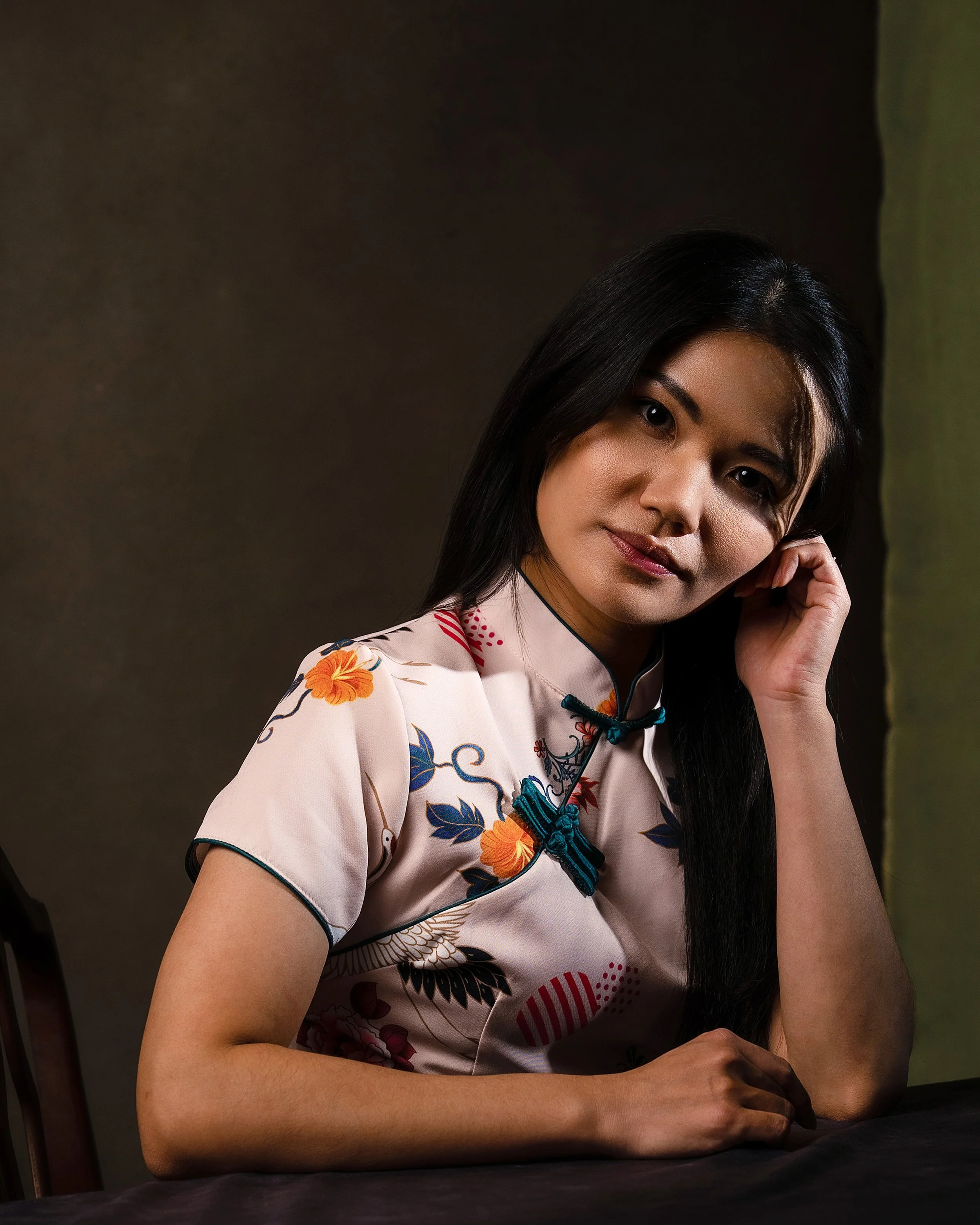 Close-up of a woman with long black hair wearing a light-colored floral patterned shirt, sitting at a table with her hand near her face against a dark background.