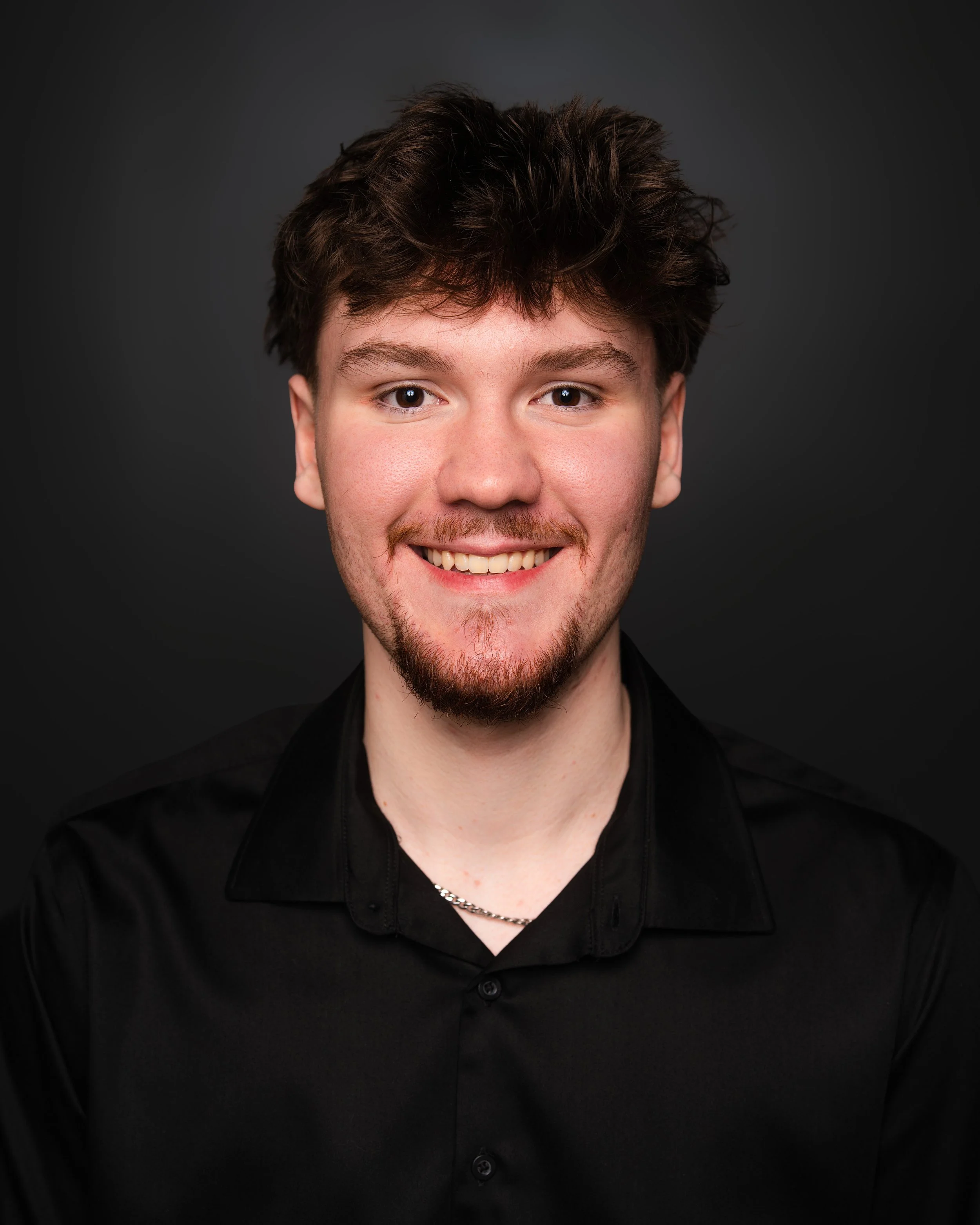 Close-up of a smiling young man with dark brown hair, light skin, wearing a black shirt and silver chain, against a dark background.