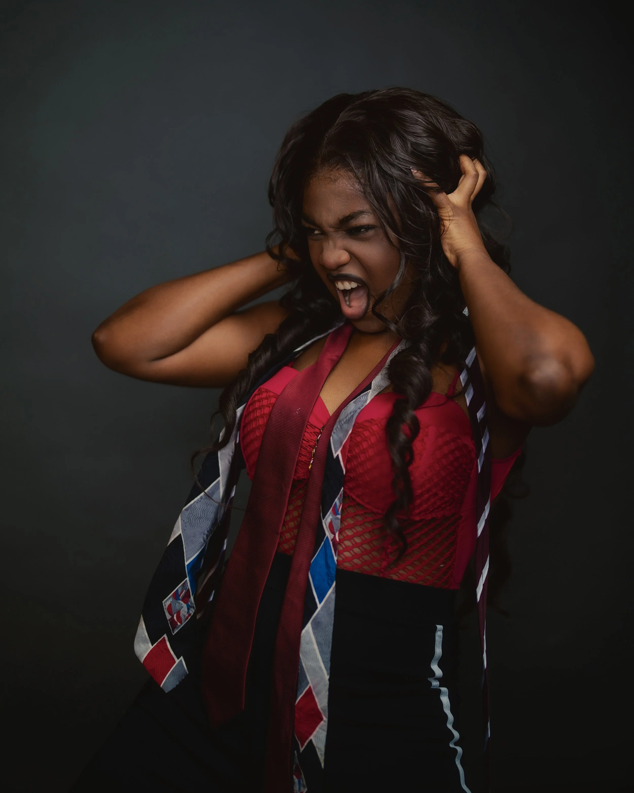 A woman with long curly black hair, wearing a red sports bra and a jacket with a colorful diamond pattern, holding her head and screaming, against a dark gray background.