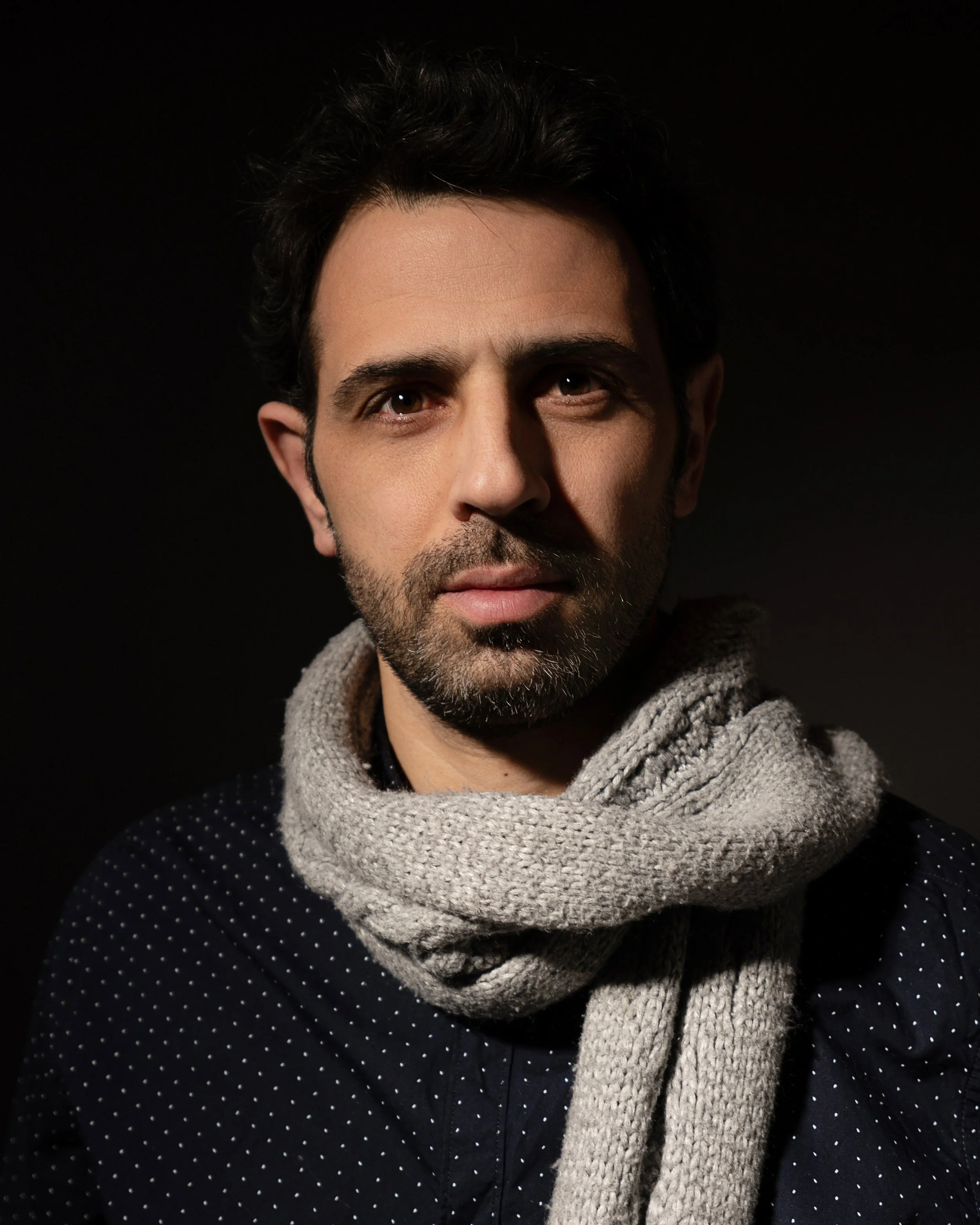 Close-up of a man with short black hair, light skin, and subtle beard, looking directly at the camera, wearing a cozy gray knit scarf around his neck and a navy blue sweater with small white polka dots, set against a black background.