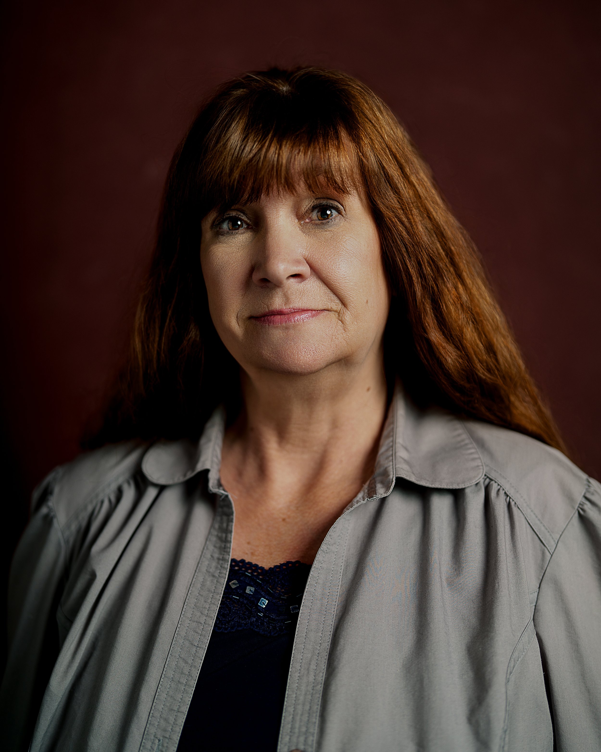 Headshot of a woman with red-brown hair in a black blouse and grey button up shirt in front of a red backdrop