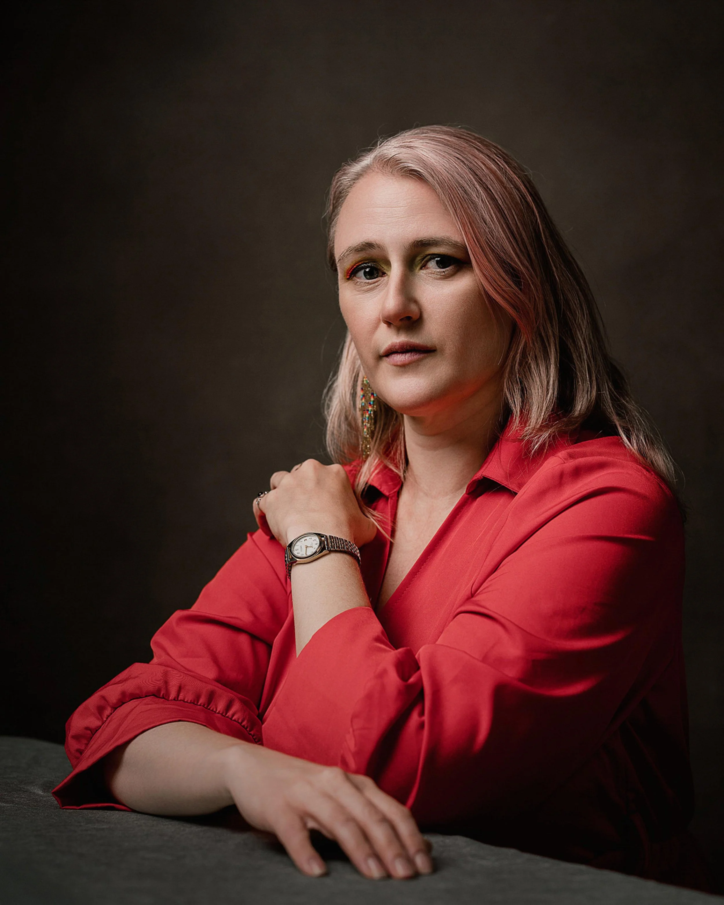 A woman with light brown hair and makeup, wearing a red long-sleeve shirt, sitting on a dark surface, with her left hand resting on the surface and her right arm crossed over her chest, wearing a watch and earrings, against a dark plain background.
