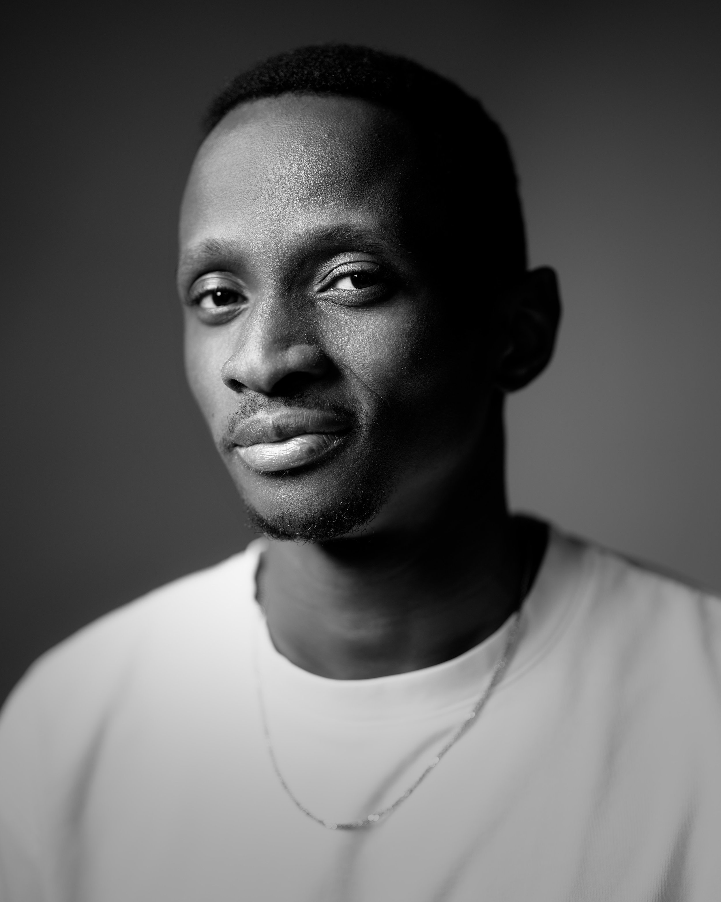 Black and white portrait of a young African American man with short natural hair, wearing a light-colored shirt and a thin chain necklace, looking at the camera with a slight smile, against a plain background.