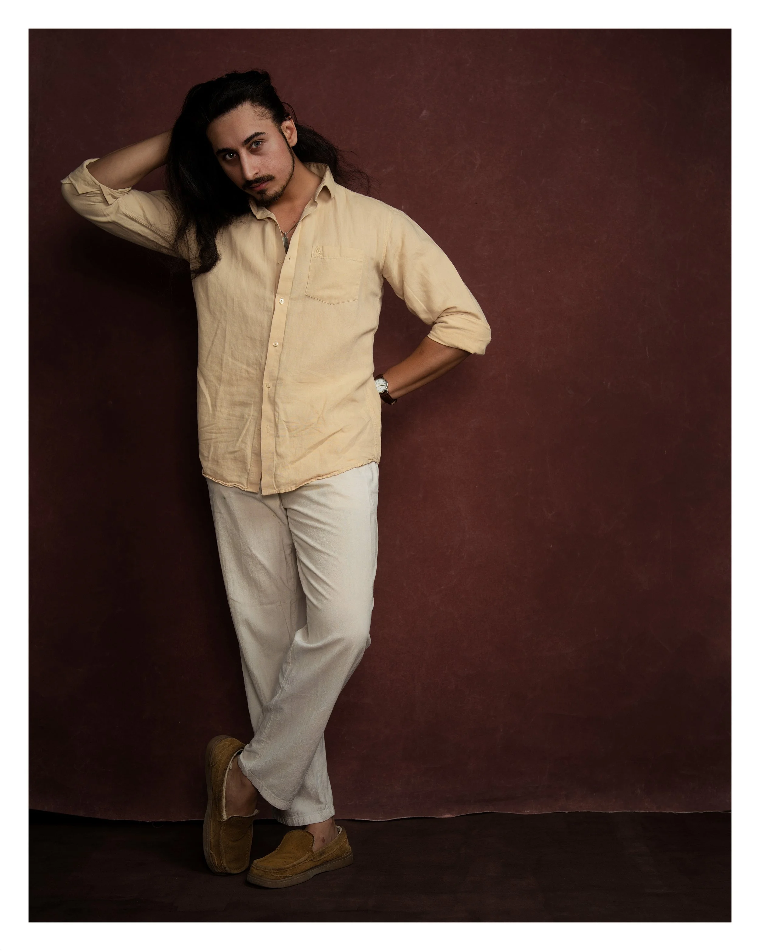 A man with long black hair, a mustache, and a beard posing against a plain maroon background. He is wearing a beige button-up shirt with the sleeves rolled up, cream-colored pants, and brown shoes. His left hand is resting on his hip, and his right h