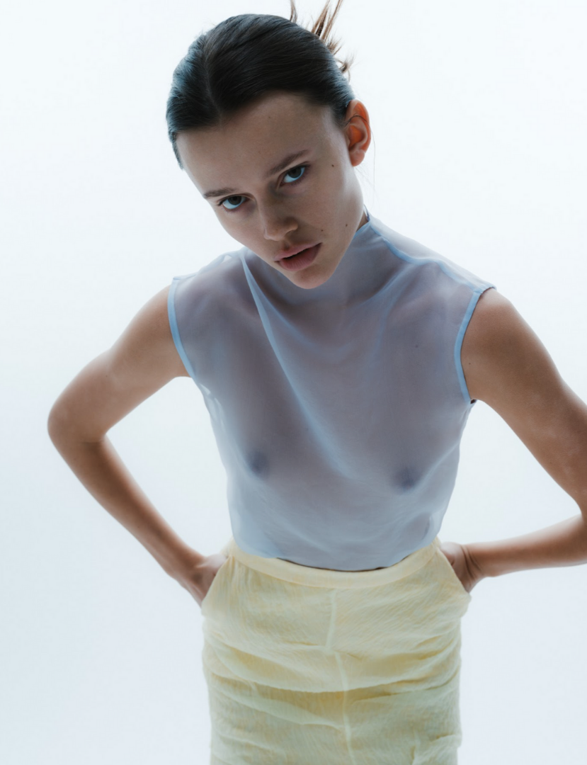 A young woman with dark hair styled in a sleek bun, wearing a transparent light blue sleeveless top and yellow shorts, standing with her hands on her hips against a plain, light background.