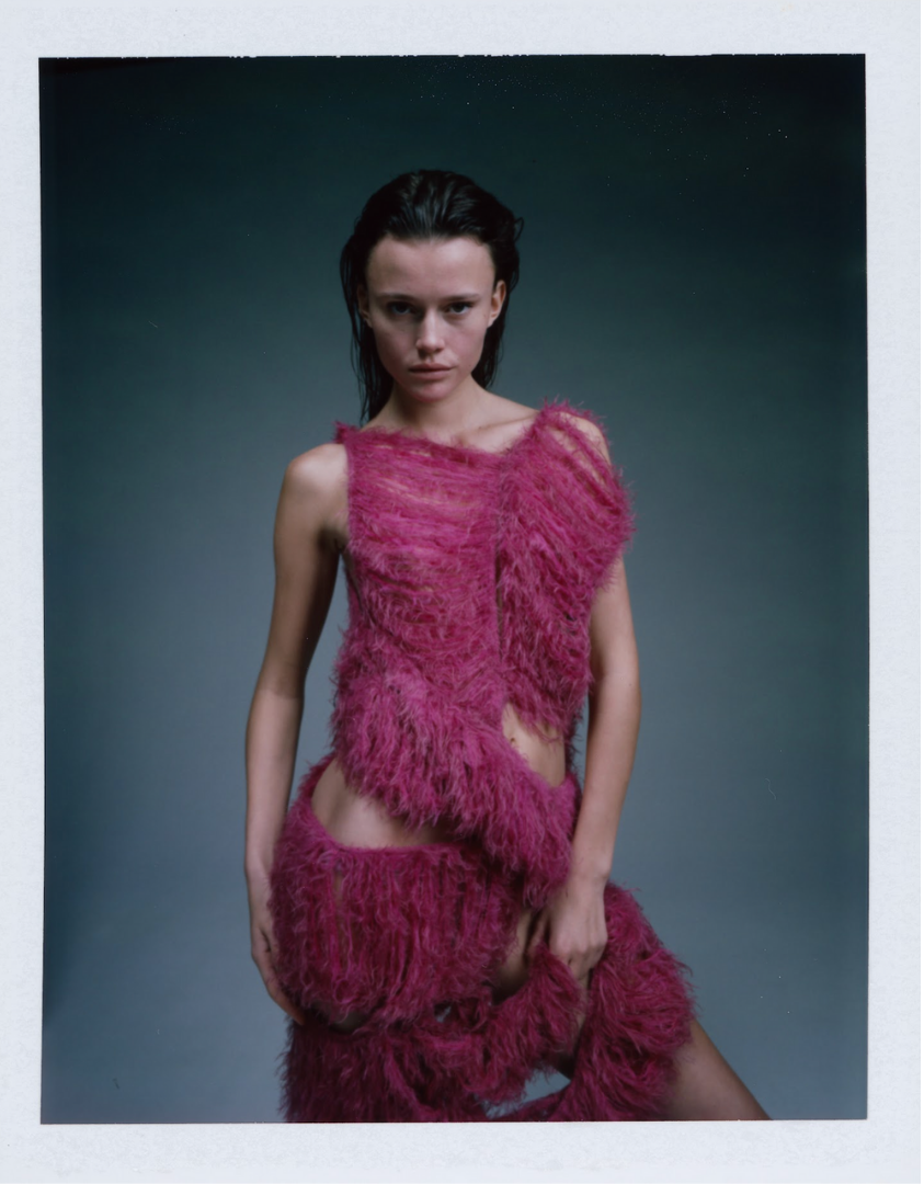 Young woman with wet hair wearing a pink, fluffy, feathered outfit against a dark gray background.