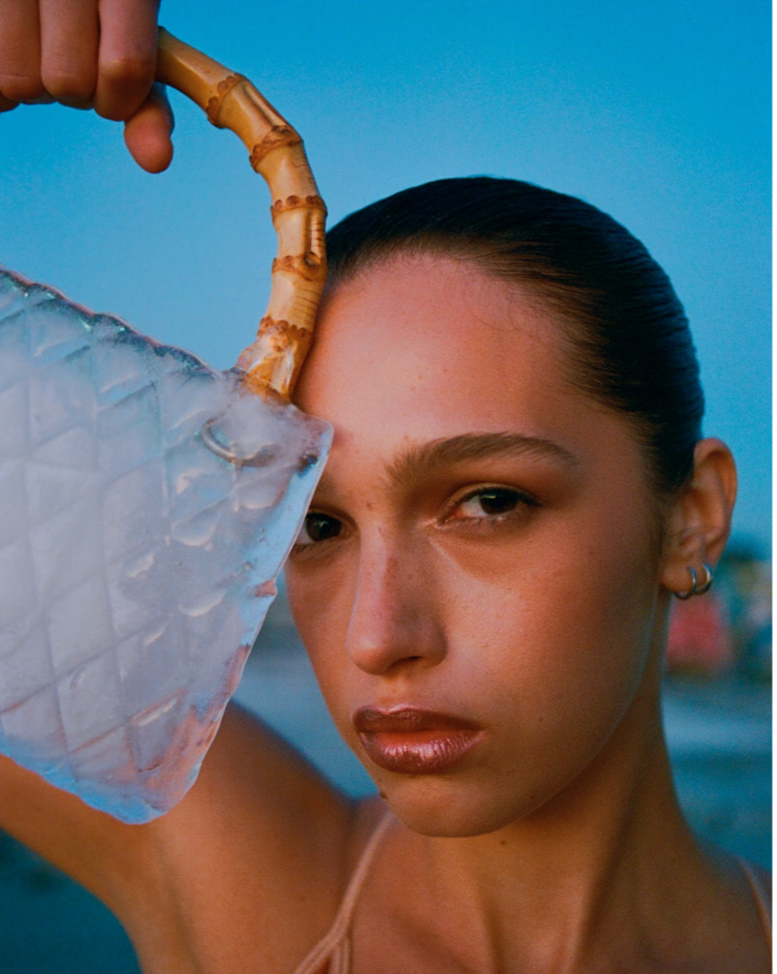 A woman with styled hair and hoop earrings holding a large seashell and a piece of ice, with a ocean or water in the background.