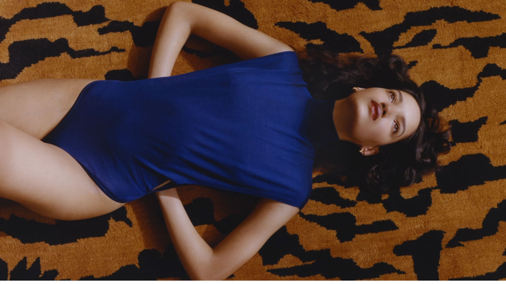 Corinne Murray lying on an orange and black patterned rug, wearing an Alaia blue bodysuit, with her arms resting behind her head, gazing upward.