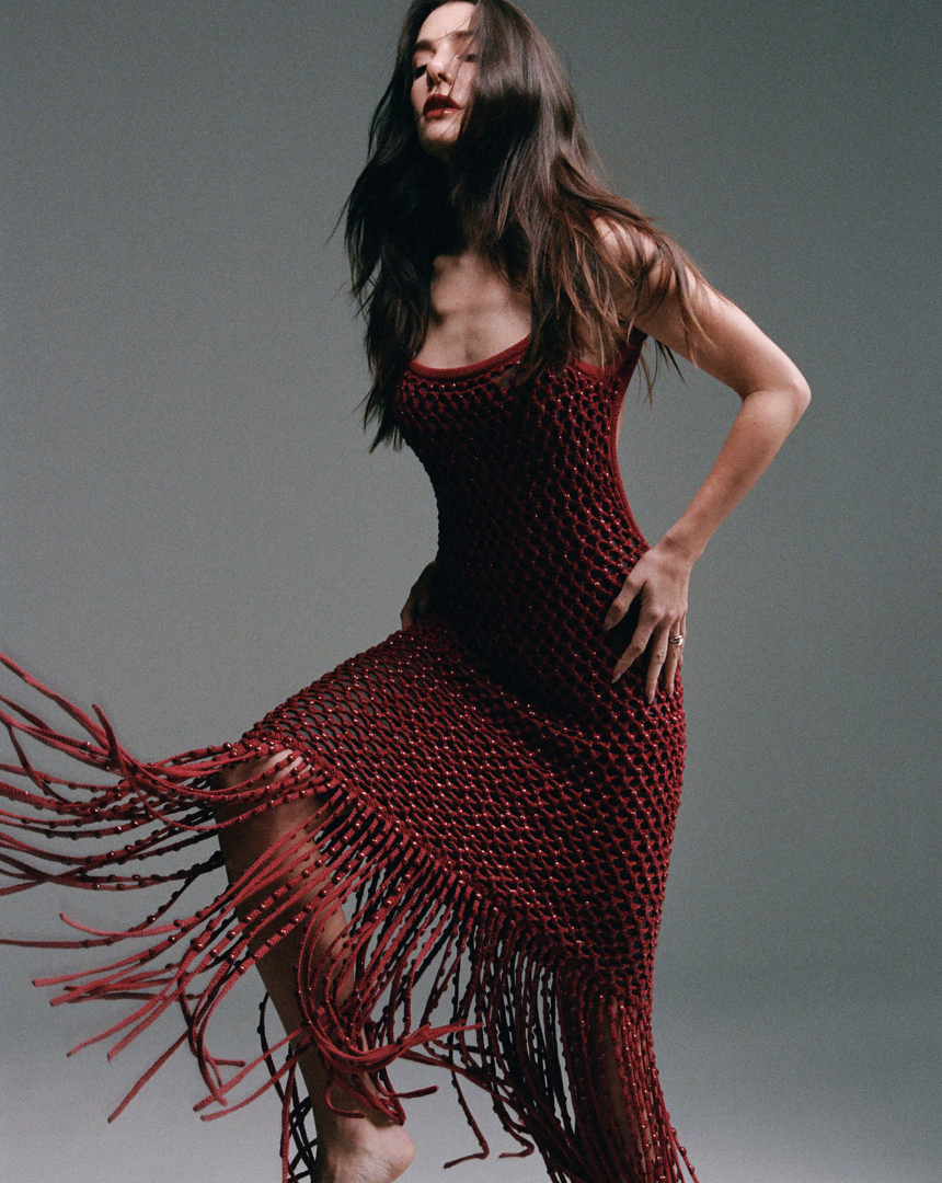 Valentina Ferrer wearing a fitted red diotima crochet dress with fringe details, posing against a gray background.
