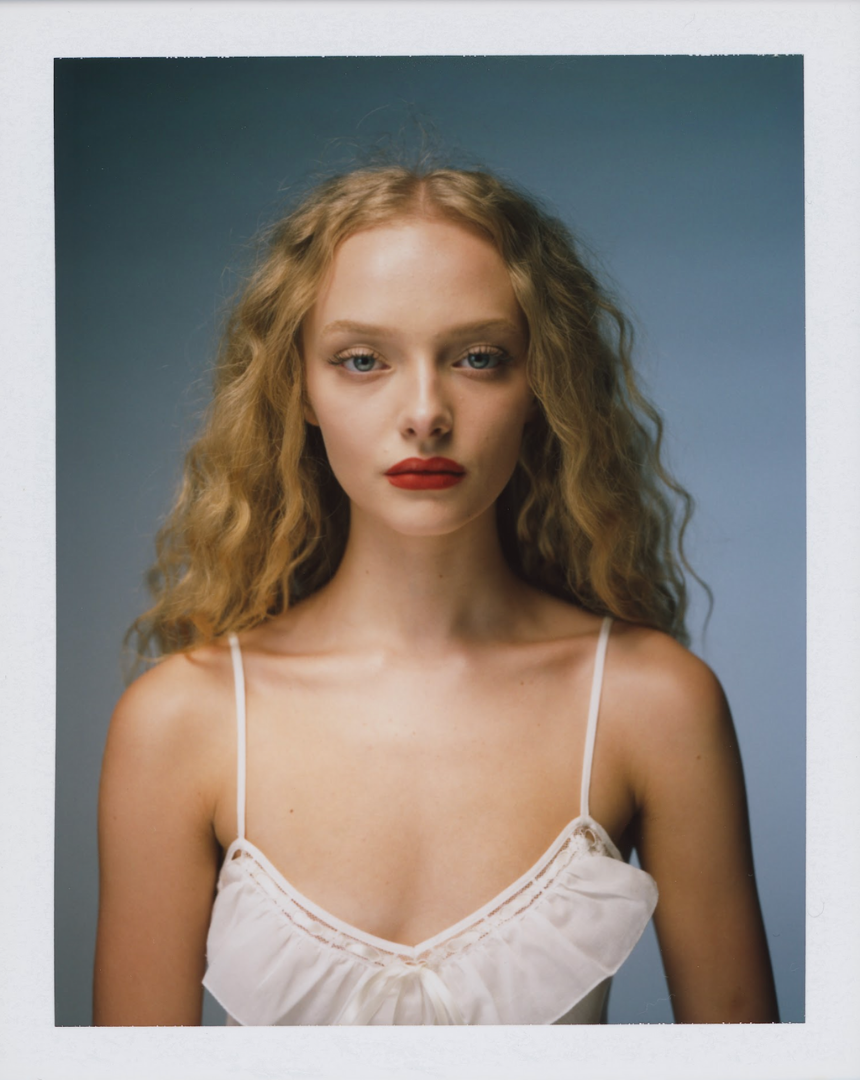 Portrait of a Amaiah Miller with curly blonde hair, wearing a white spaghetti strap top, with natural makeup and red lipstick, against a blue background.
