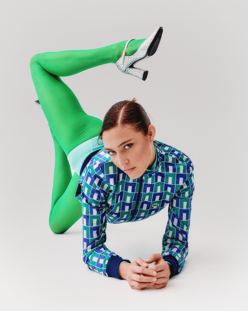 Woman lying on the floor in a crawling pose, wearing a colorful geometric patterned jacket, green tights, and glittery high heels, against a plain white background. Bottega Venetta Silver Heels