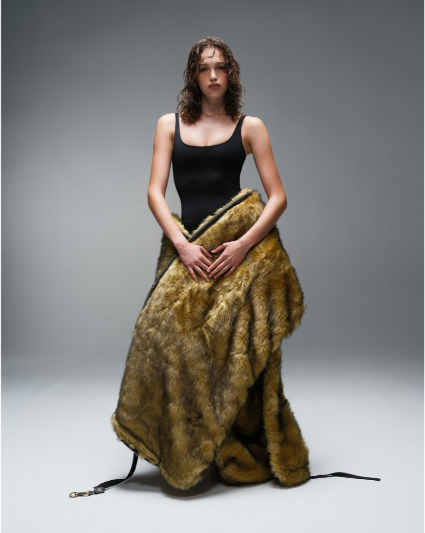 A woman with wavy brown hair standing against a plain gray background, wearing a fitted black top and a large, brown fur coat draped around her waist.