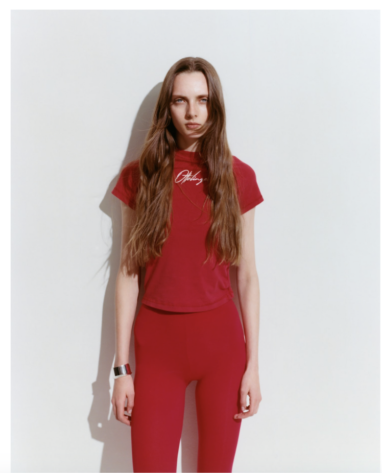Young woman with long brown hair wearing a red ottolinger t-shirt and matching red pants standing against a plain white wall.