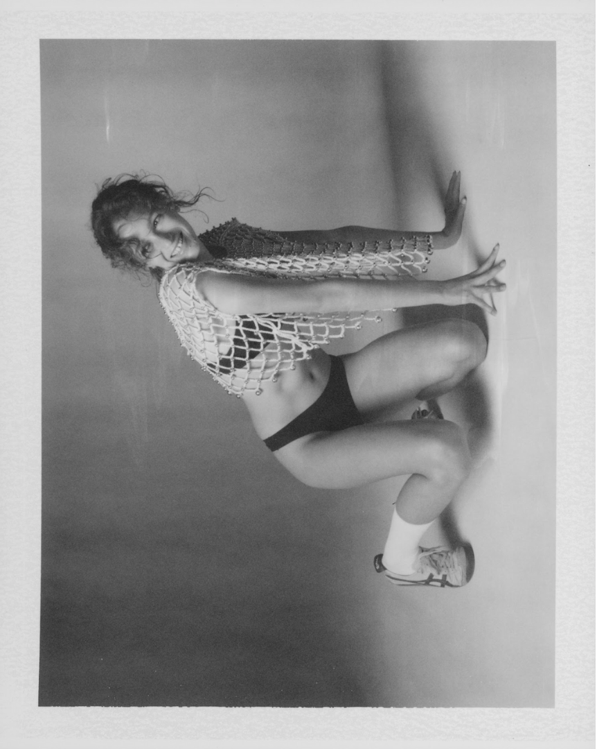 Woman in black shorts and sneakers squatting against a plain wall, wearing a crochet top, smiling, black and white photo.