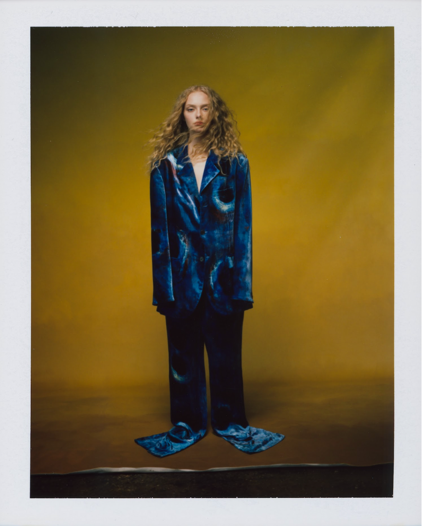 Amaiah Miller in Marni with long curly hair wearing a blue tie-dye suit standing in front of a brown background.
