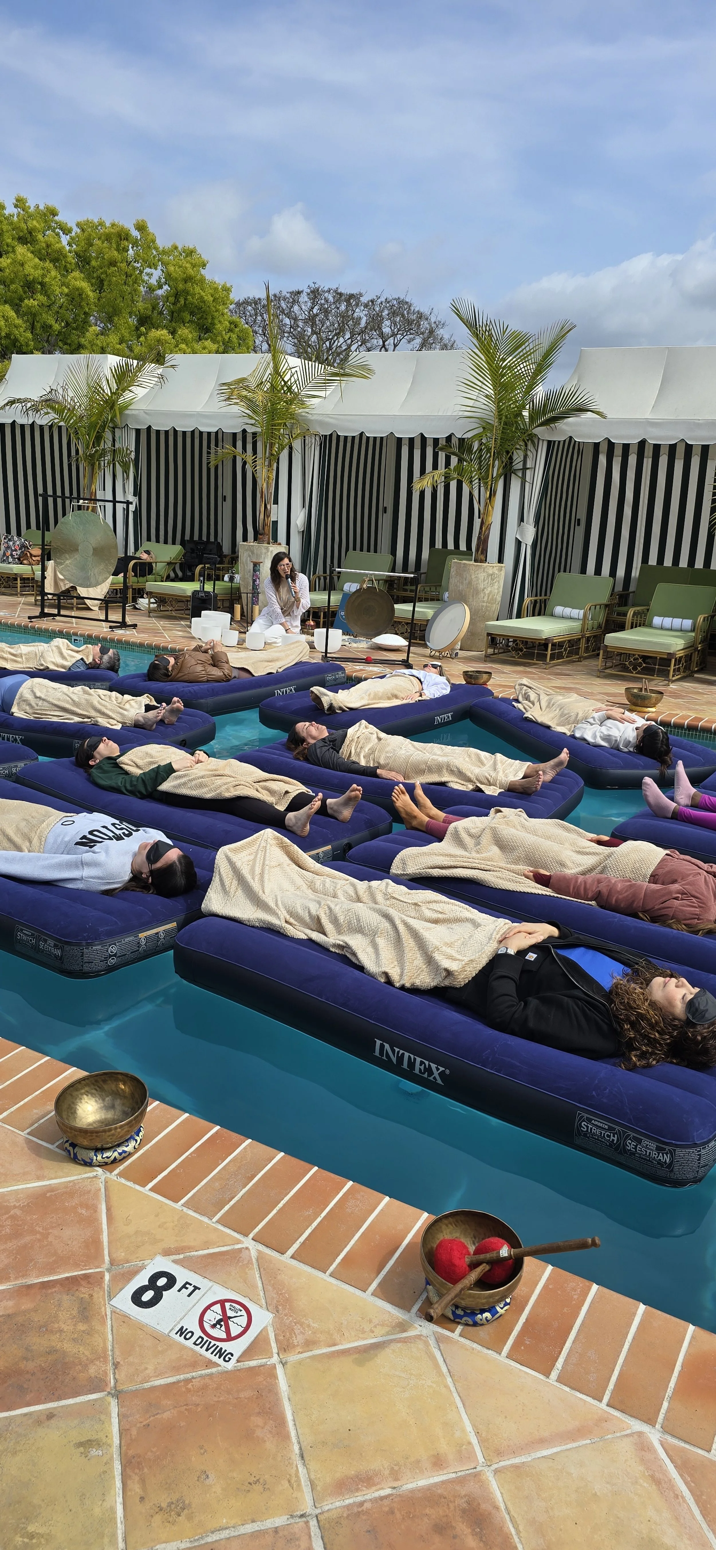 People participating in a sound healing or relaxation session by a poolside, lying on inflatable beds, with some blankets covering them, under a partly cloudy sky with green trees in the background.