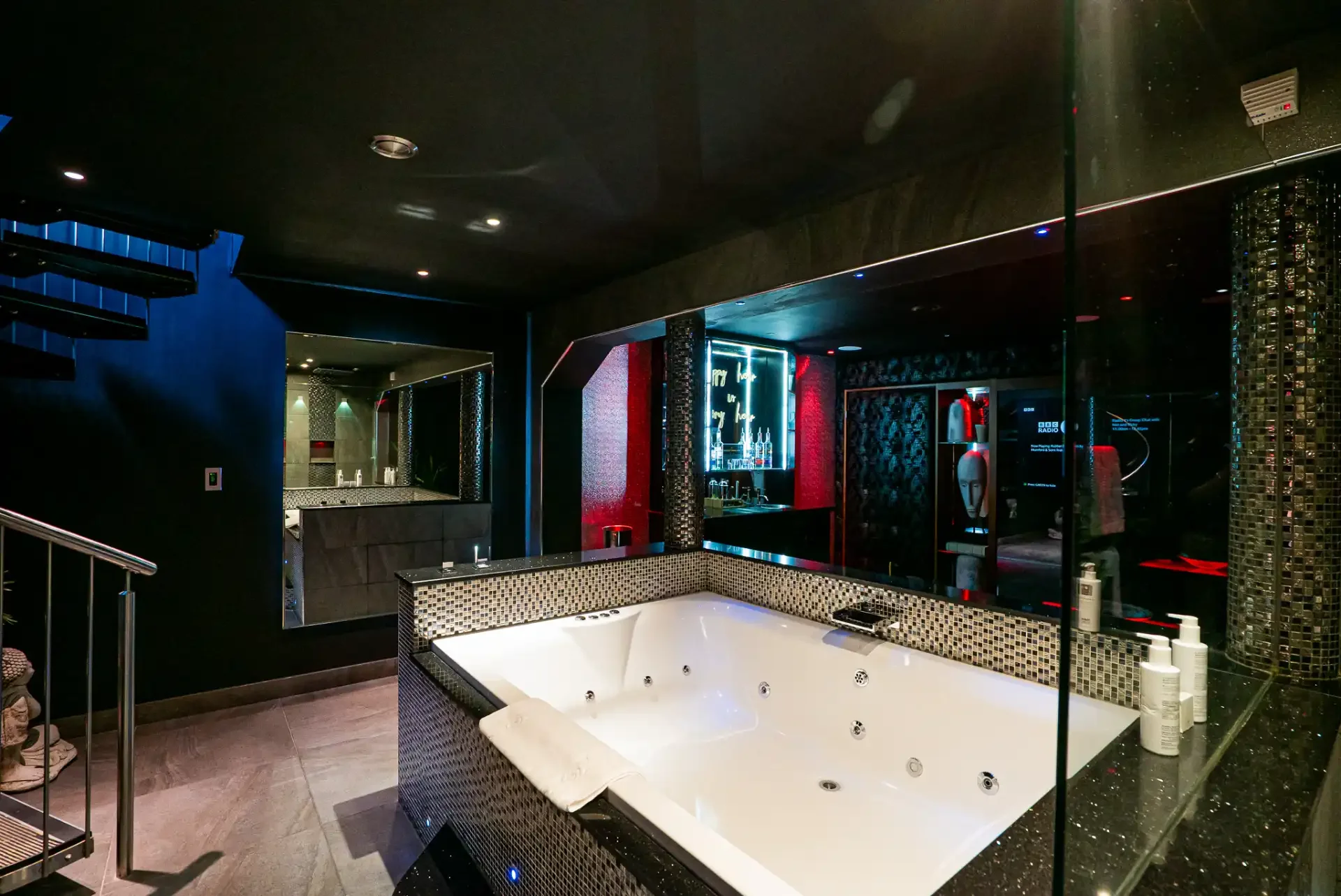Indoor spa area with a large jacuzzi tub, black walls, and reflective surfaces, decorated with colorful lights and toiletries on the edge of the tub.