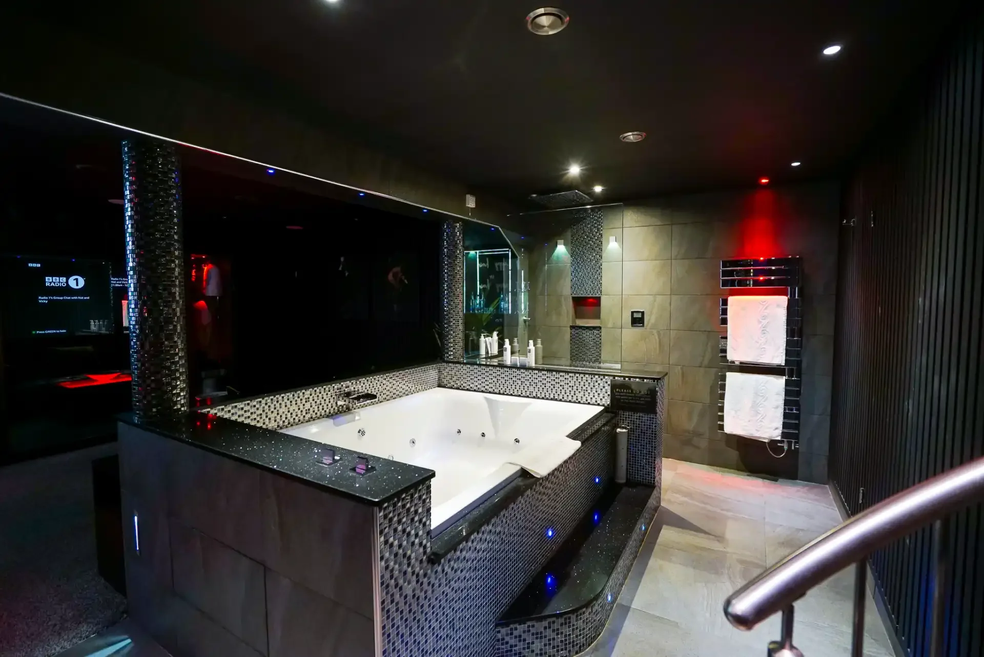 A modern indoor spa room with a large white jacuzzi tub surrounded by black and silver mosaic tiles. The room has dim lighting, a mirror, and black beadboard walls, with folded white towels on a black towel rack and various spa products on the ledge.