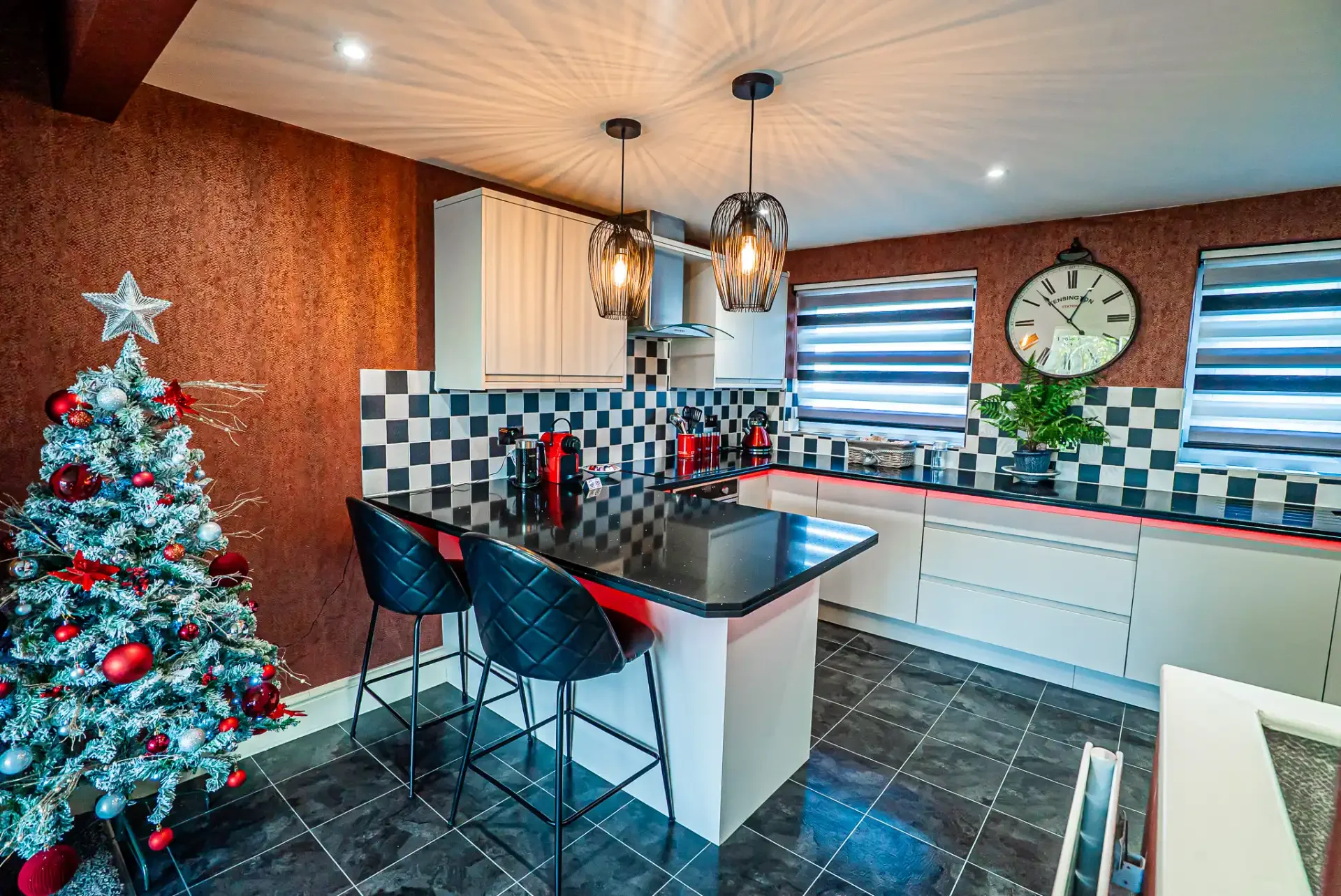 Modern kitchen decorated for Christmas with a Christmas tree adorned with red and silver ornaments, black and white checkered backsplash, white cabinets, black countertops, two black pendant lights, a potted plant, and windows with black and white ho