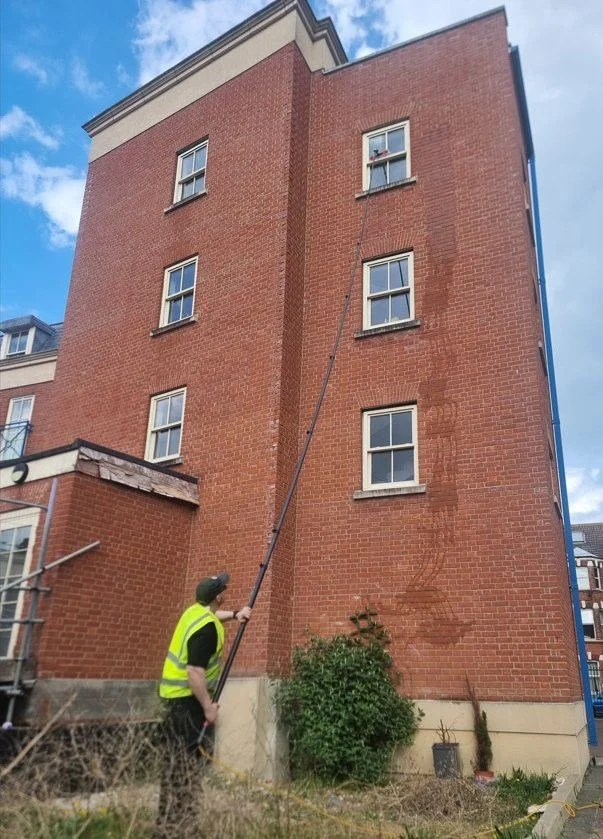 Window cleaner in high-visibility vest using a water-fed pole on a five-storey red brick building in Canterbury