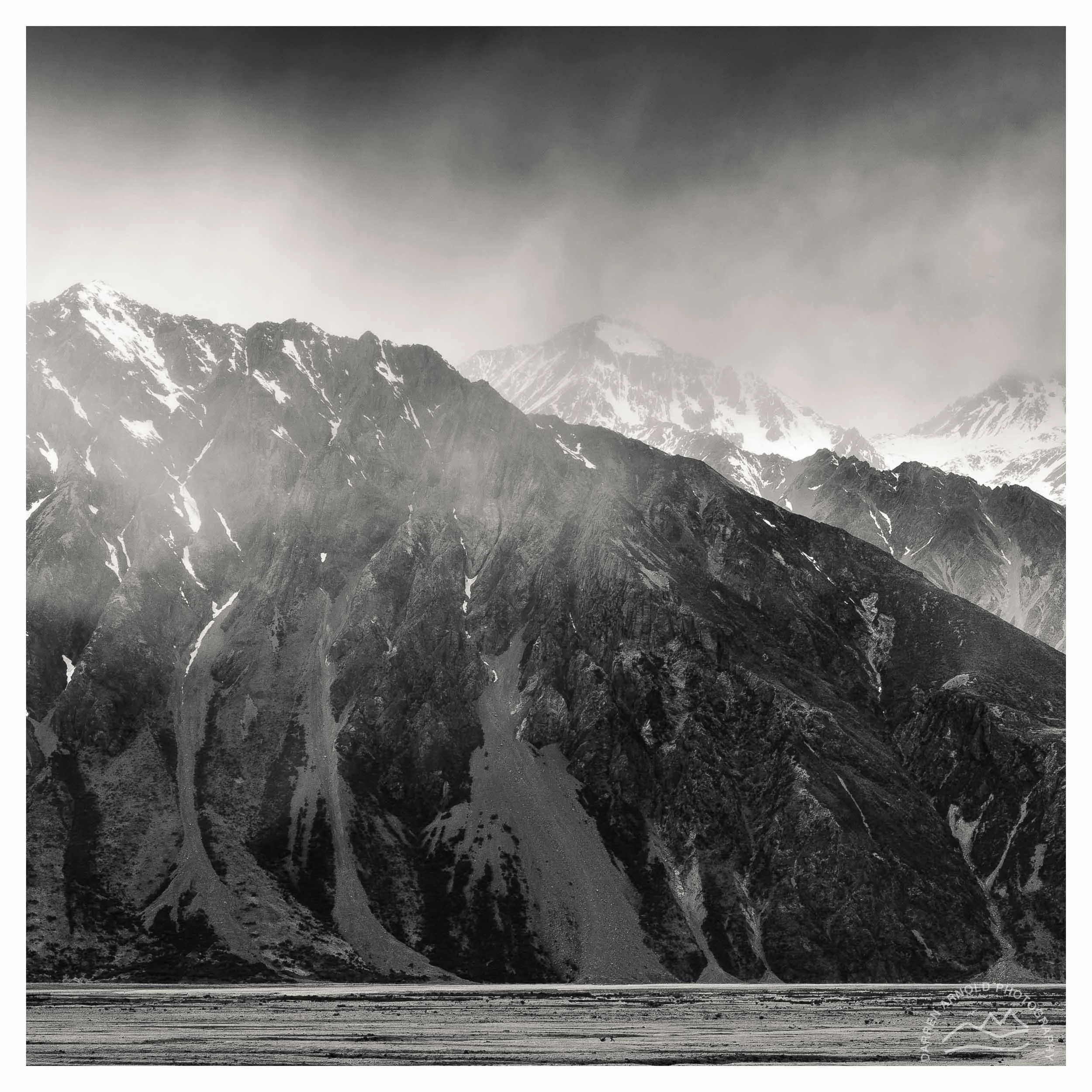 Black and white photo of a mountain range with snow-capped peaks and steep slopes, with a flat area of land at the base.