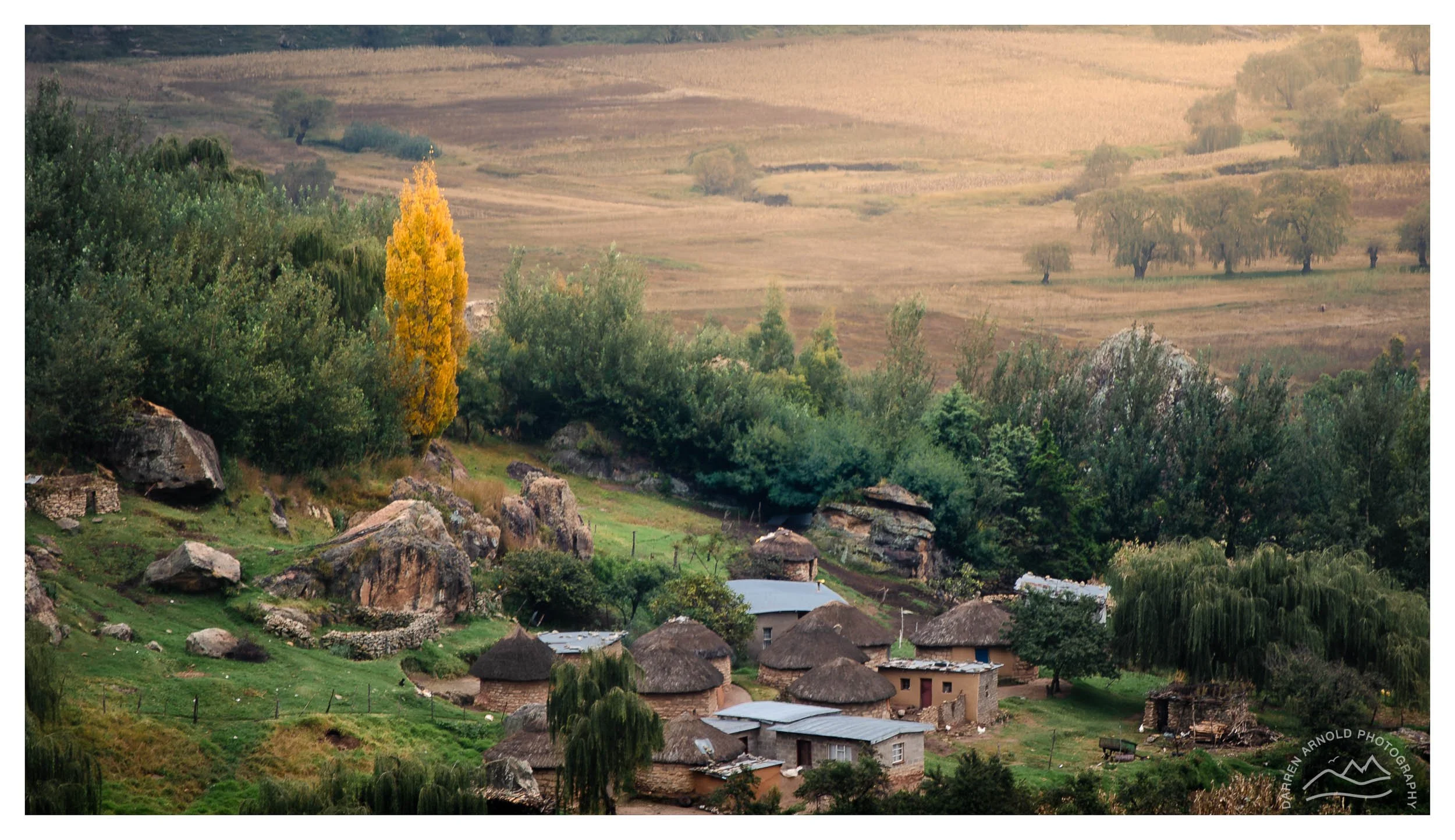 A rural village with mud and thatched-roof houses nestled among green trees and large boulders, with rolling hills and fields in the background.