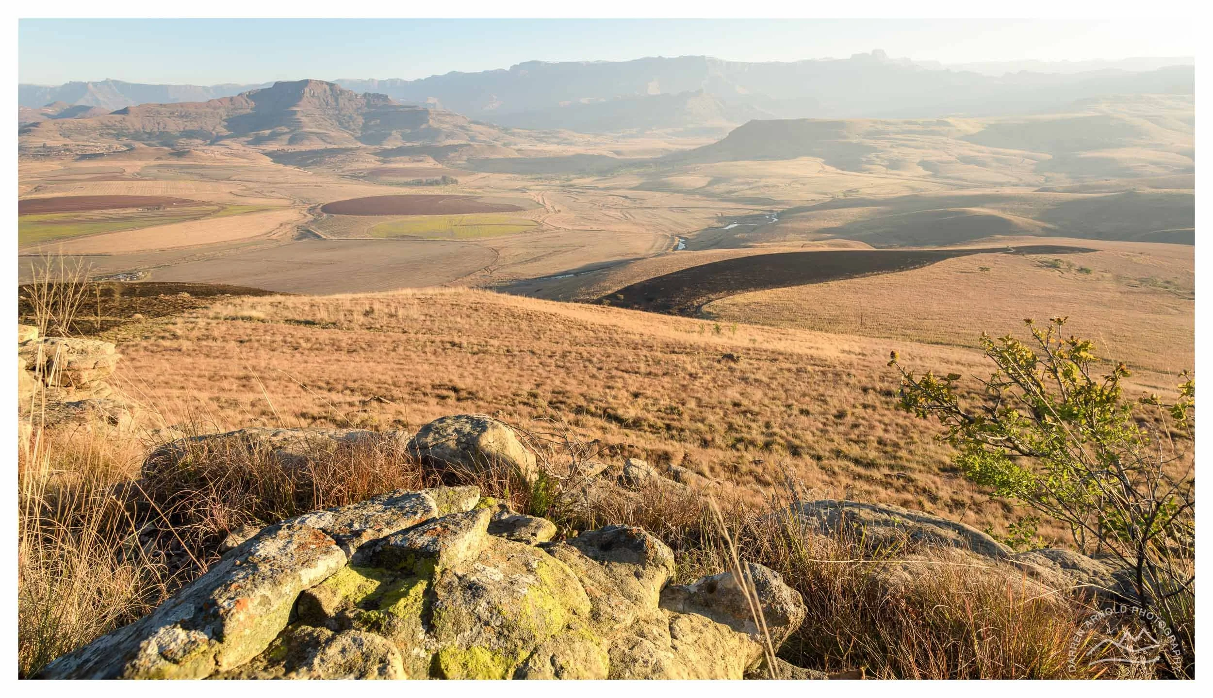 Landscape view of rolling hills, fields, and distant mountains with rocks and grass in the foreground.
