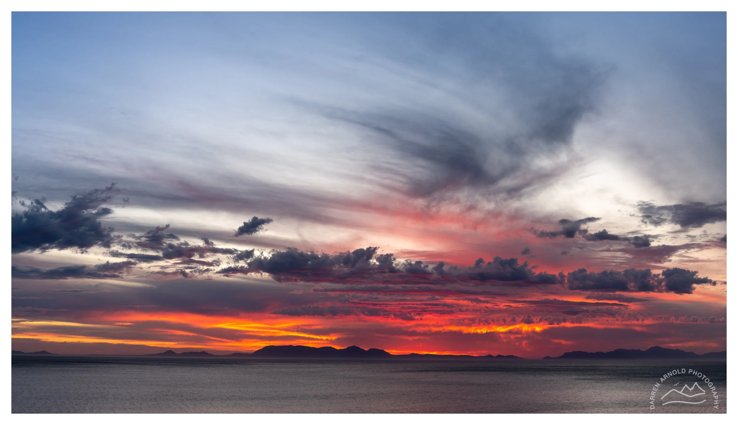 Sunset over a body of water with mountains in the distance, colorful clouds in the sky displaying shades of orange, pink, and purple.
