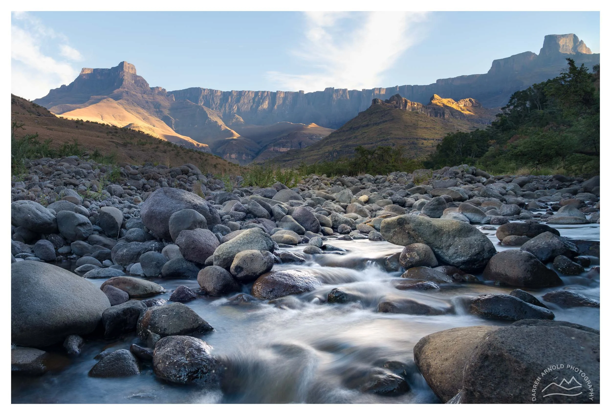 Rocky riverbed with flowing water in foreground, lush green hills and rugged mountains in background under clear blue sky.