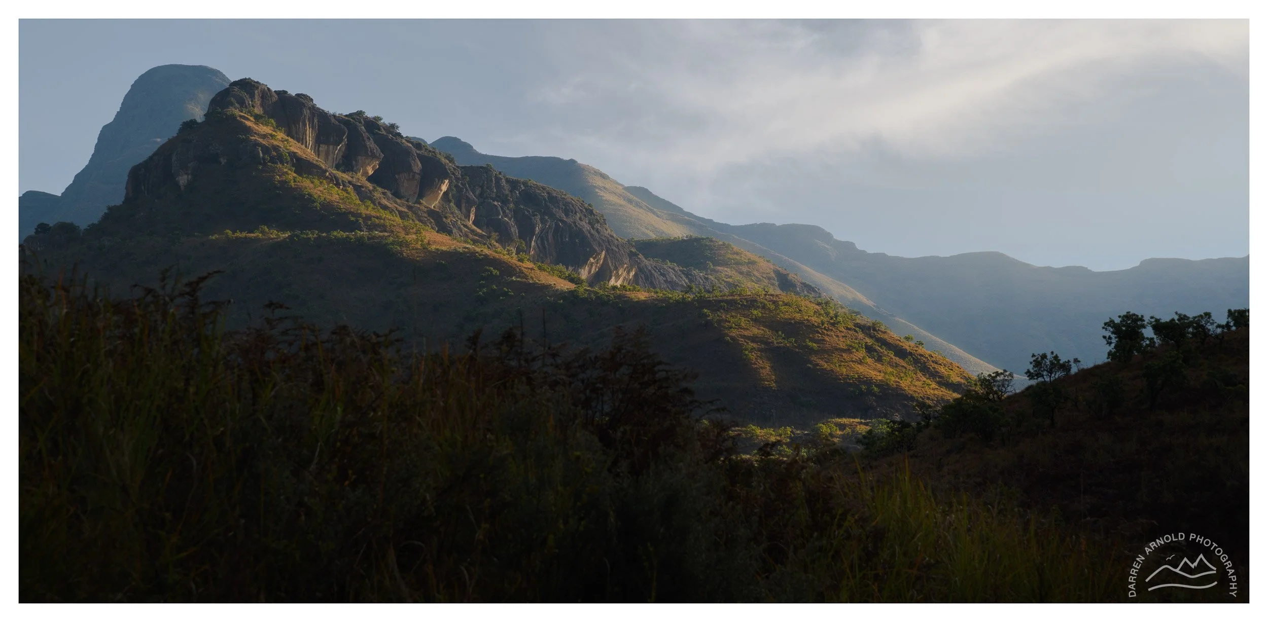 Mountain range with lush greenery and trees, under a partly cloudy sky.