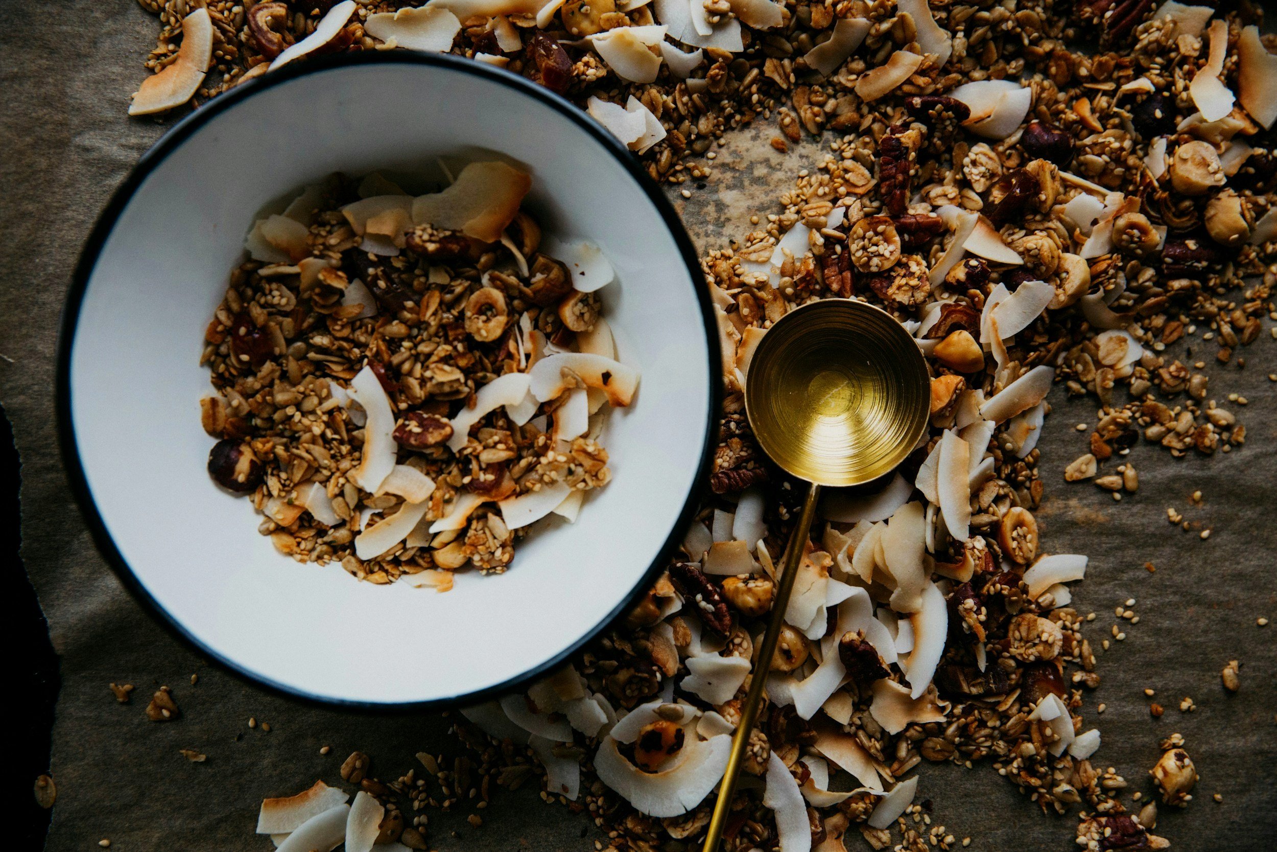 A white bowl filled with granola, coconut flakes, and nuts, with granola and coconut scattered on the surface around it, and a golden spoon resting among the scattered mixture.
