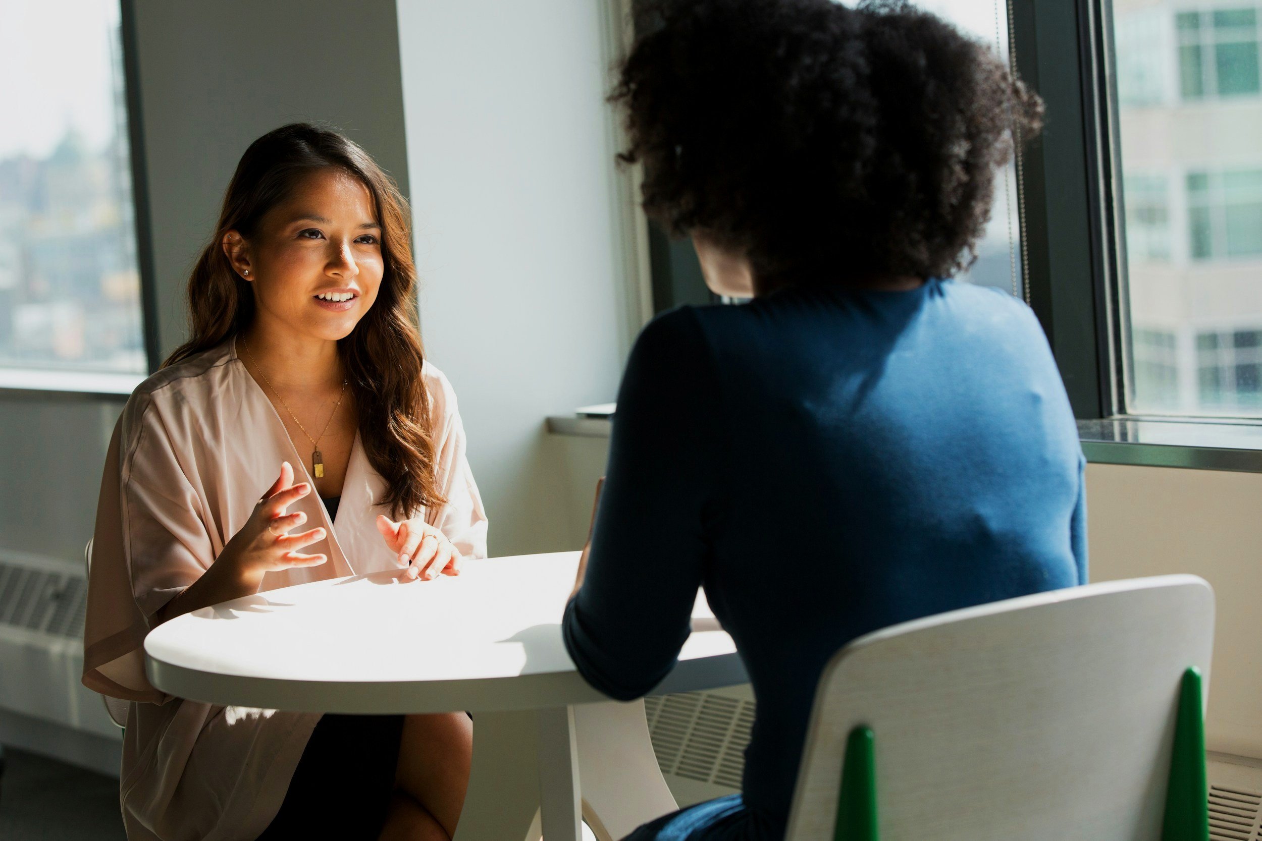 Two women having a conversation at a white table near a window in a bright room.