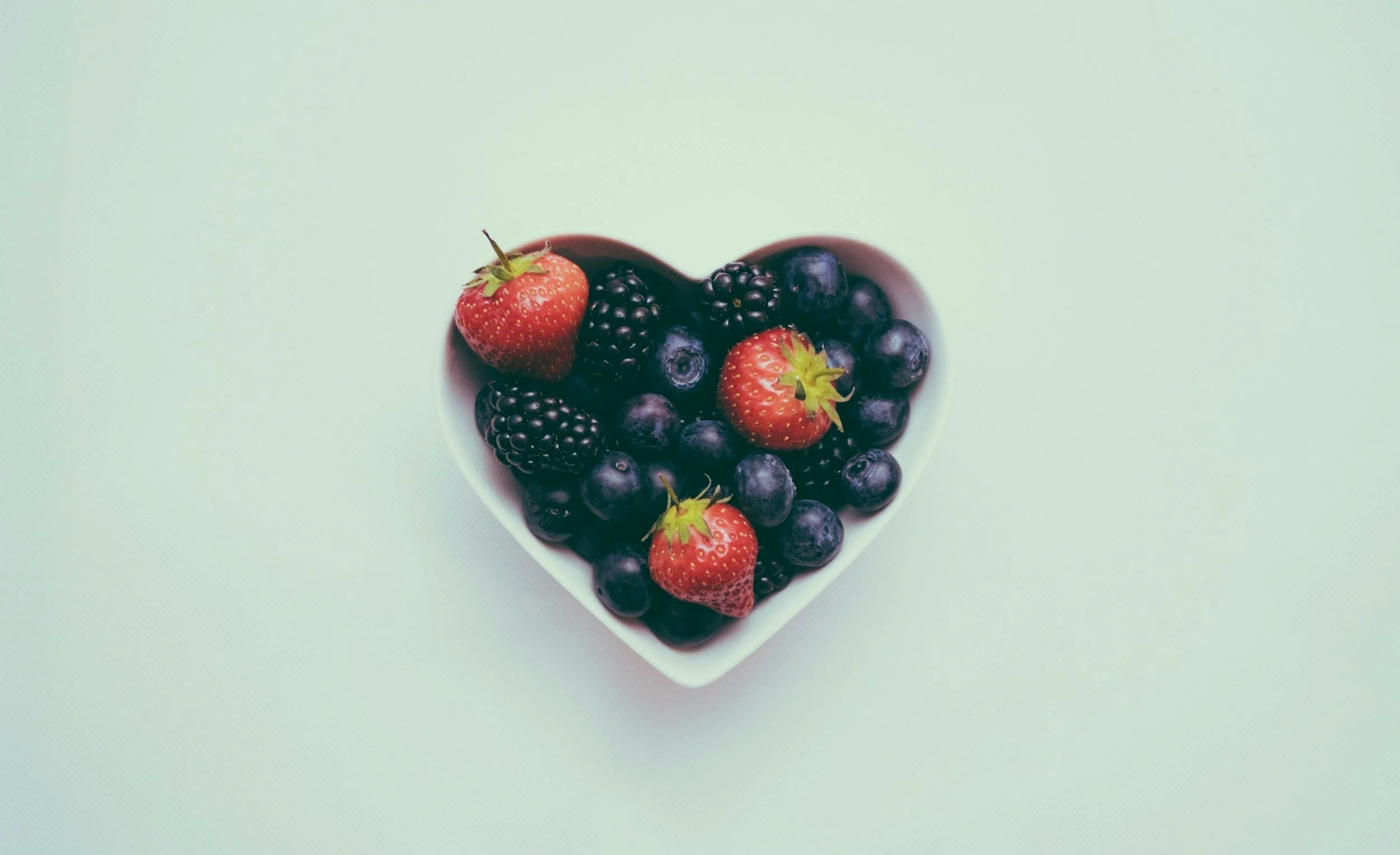 Heart-shaped bowl filled with strawberries, blackberries, and blueberries on a pastel green background.