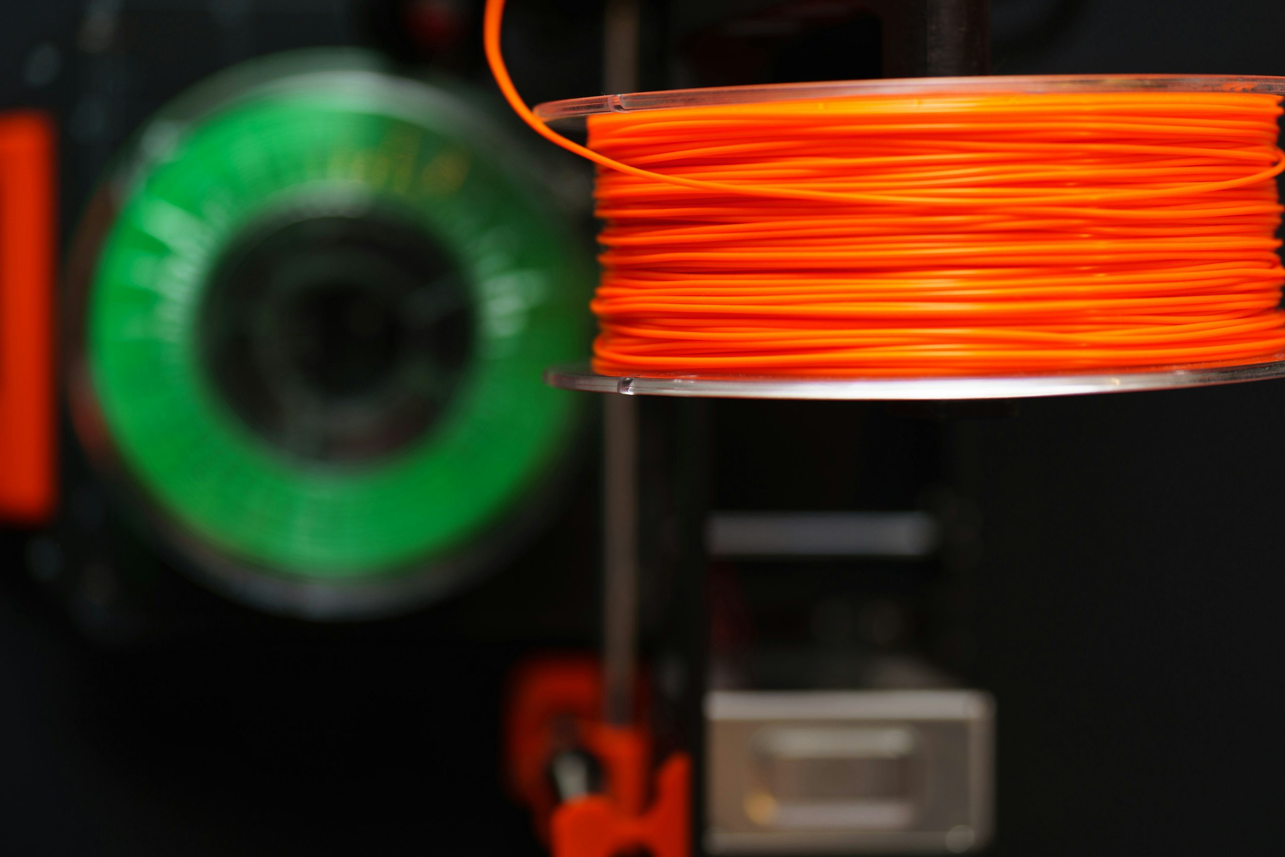 Close-up of a spool of orange filament on a 3D printer with a green component in the background.