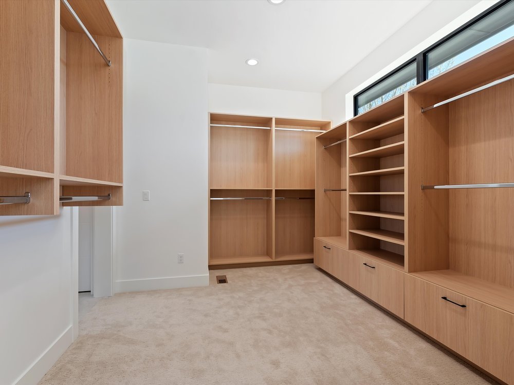 Eagle, Idaho - Eco Builders Spacious walk-in closet with wooden shelving and hanging rods, beige carpet, and a high window for natural light.