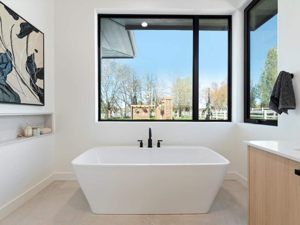 Eagle, Idaho - Eco Builders Modern bathroom with a large white freestanding bathtub, large windows with a scenic outdoor view, a towel hanging on a side wall, and abstract artwork. Minimalist design with neutral tones.